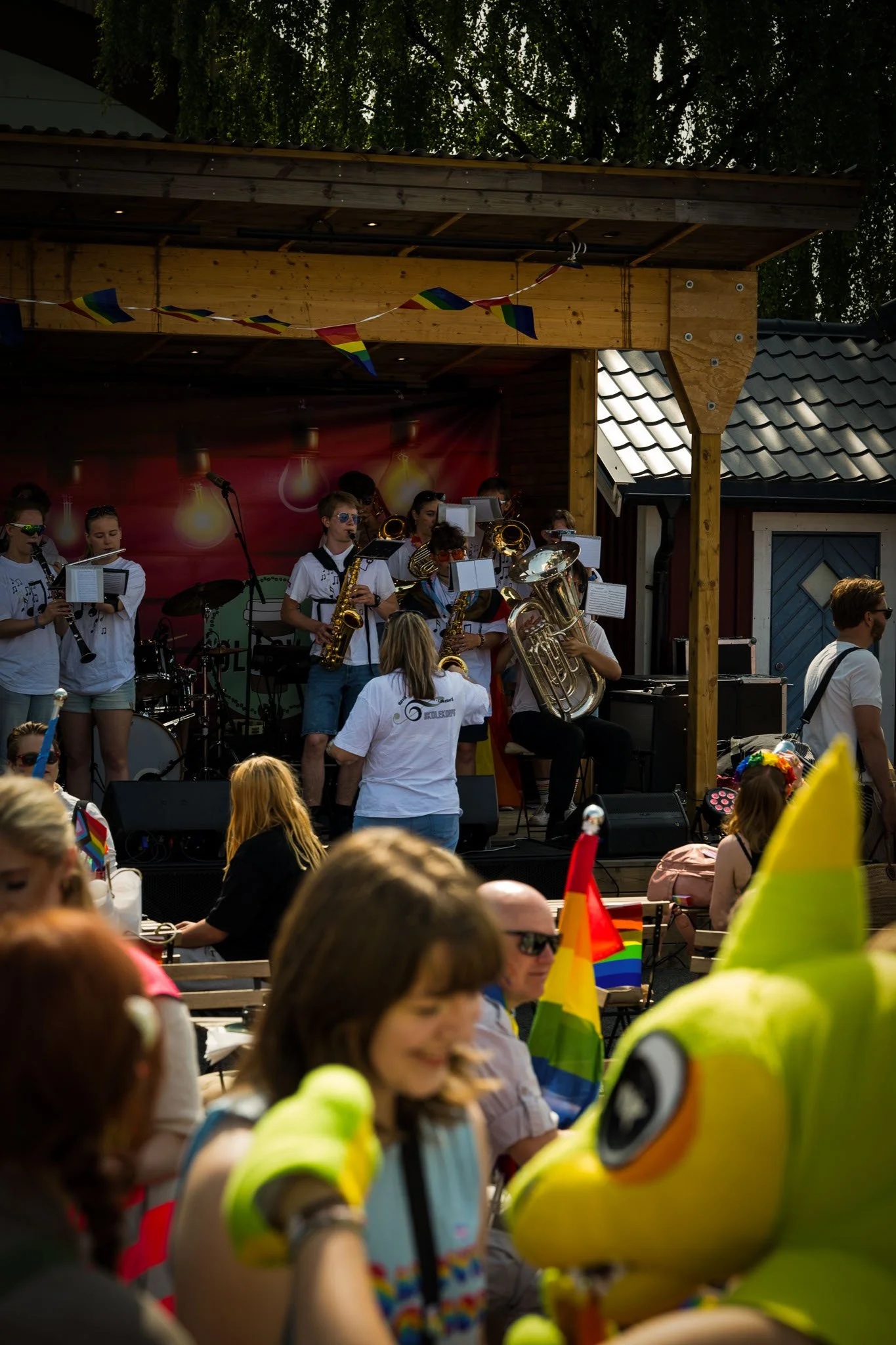 En ungdomsband spiller på et utendørs konsertevent med folk som ser på, noen med regnbueflagger og en karnevalskostyme i forgrunnen.