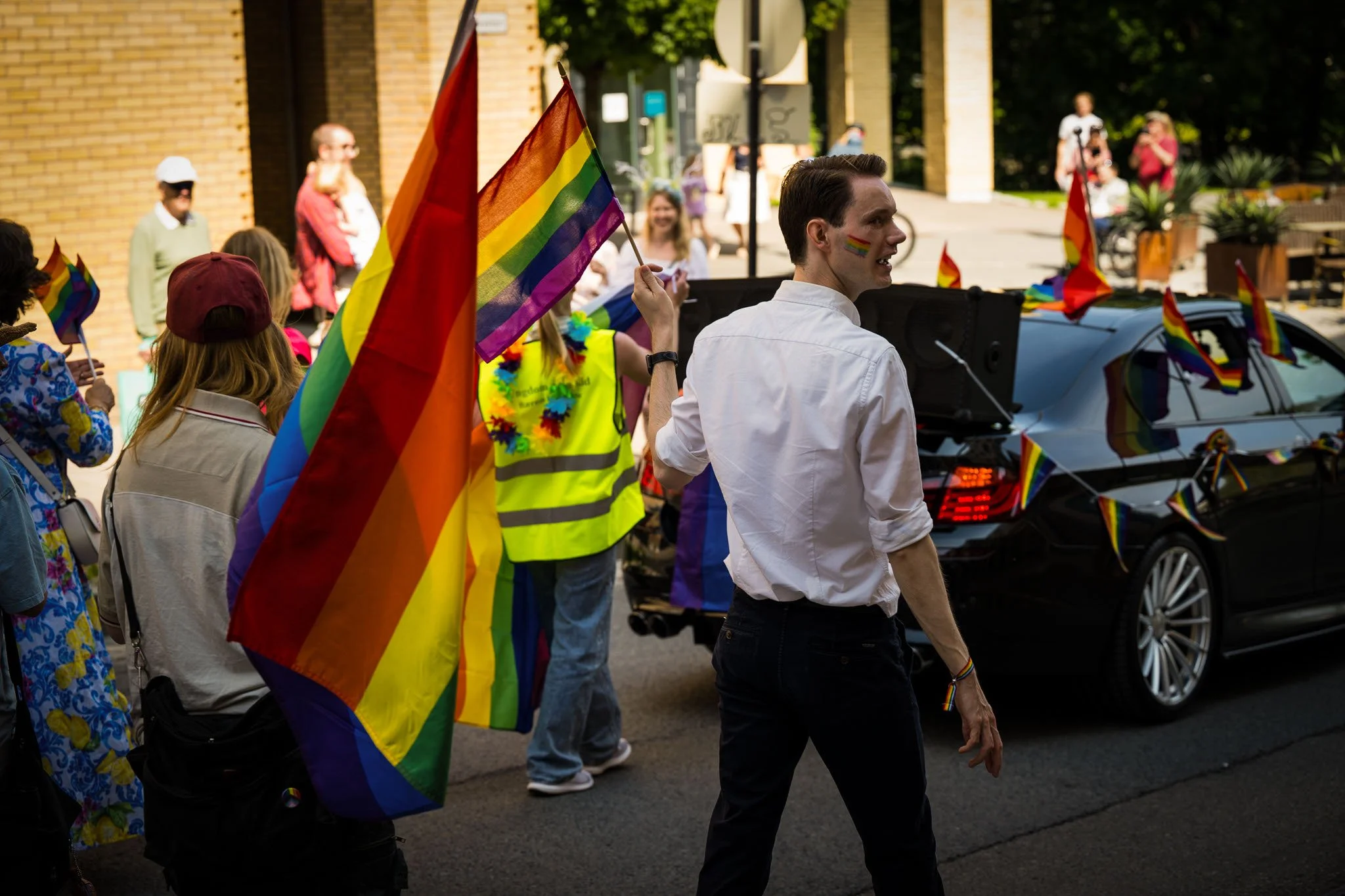 Person som deltar i en Pride-parade med regnbågflagga, omringet av andre festkledde personer, med biler dekorert med Pride-flagg i bakgrunnen.