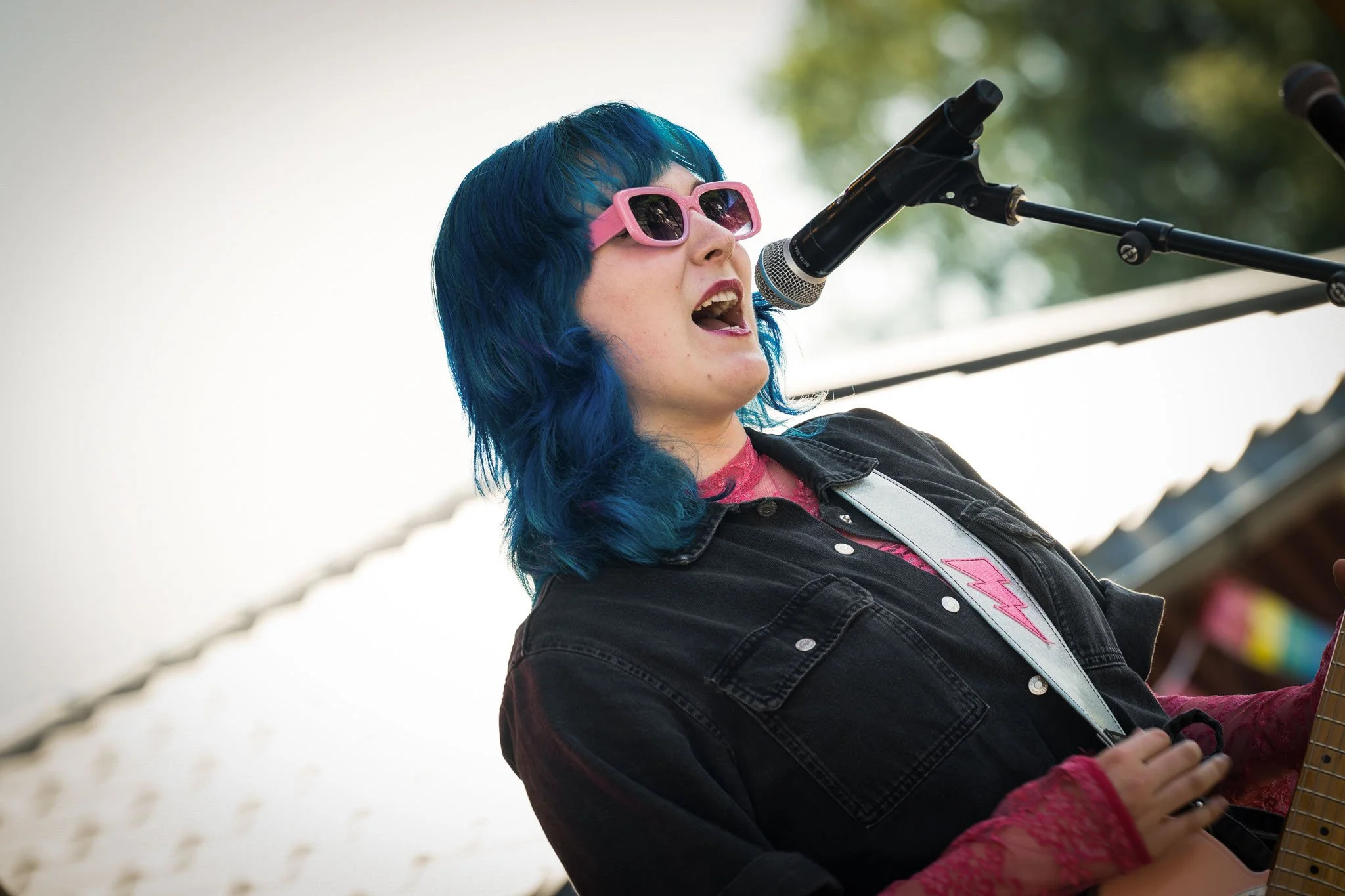 En kvinnelig musiker med blå hår og rosa solbriller synger inn i en mikrofon mens hun spiller gitar, utendørs.