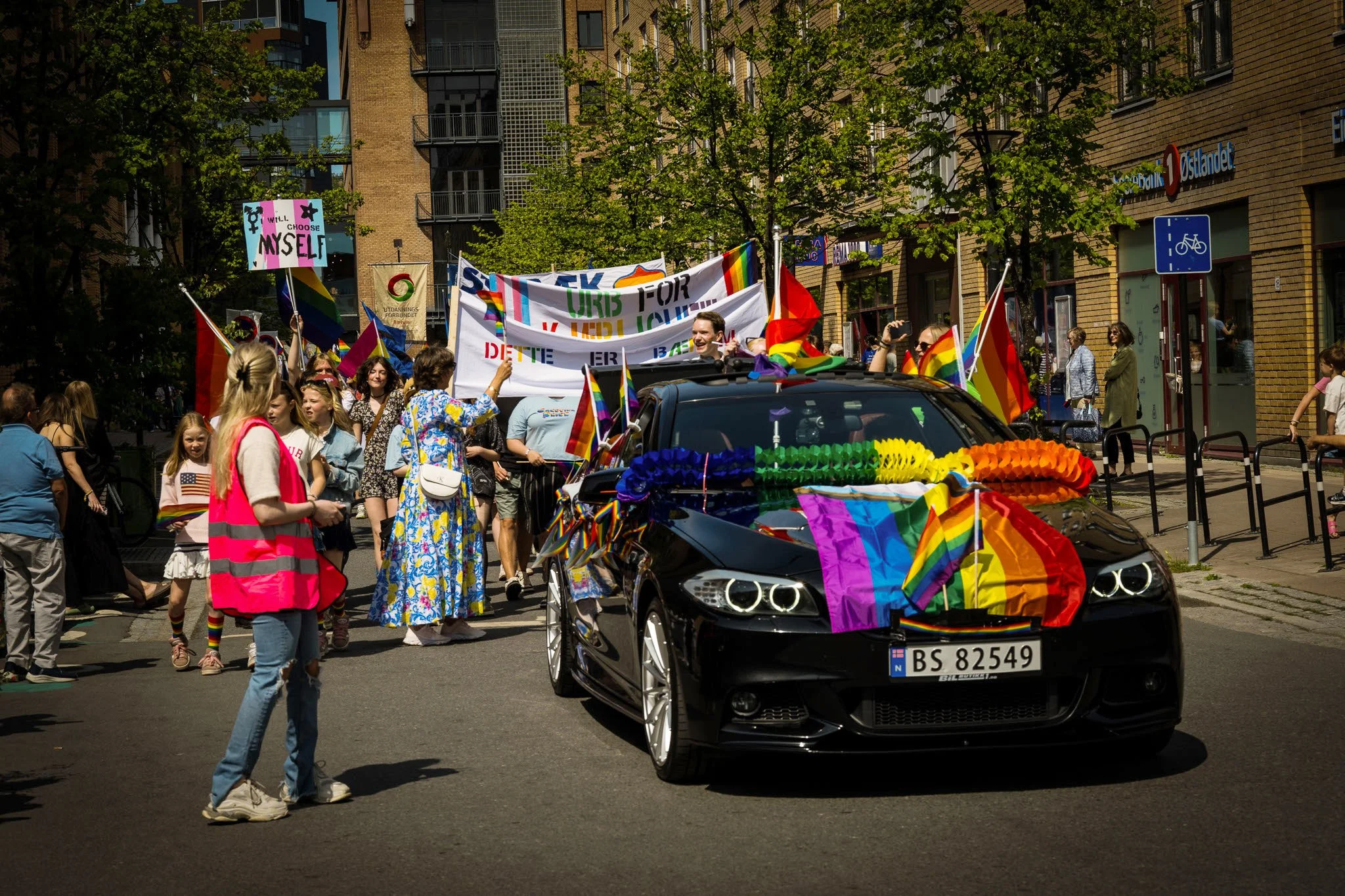 En svart bil dekorert med regnbueflagger og blomster i en parade eller feiring, med en gruppe mennesker og flagg som bærer regnbue- og pride-flagg, der mange mennesker feirer i bakgrunnen.
