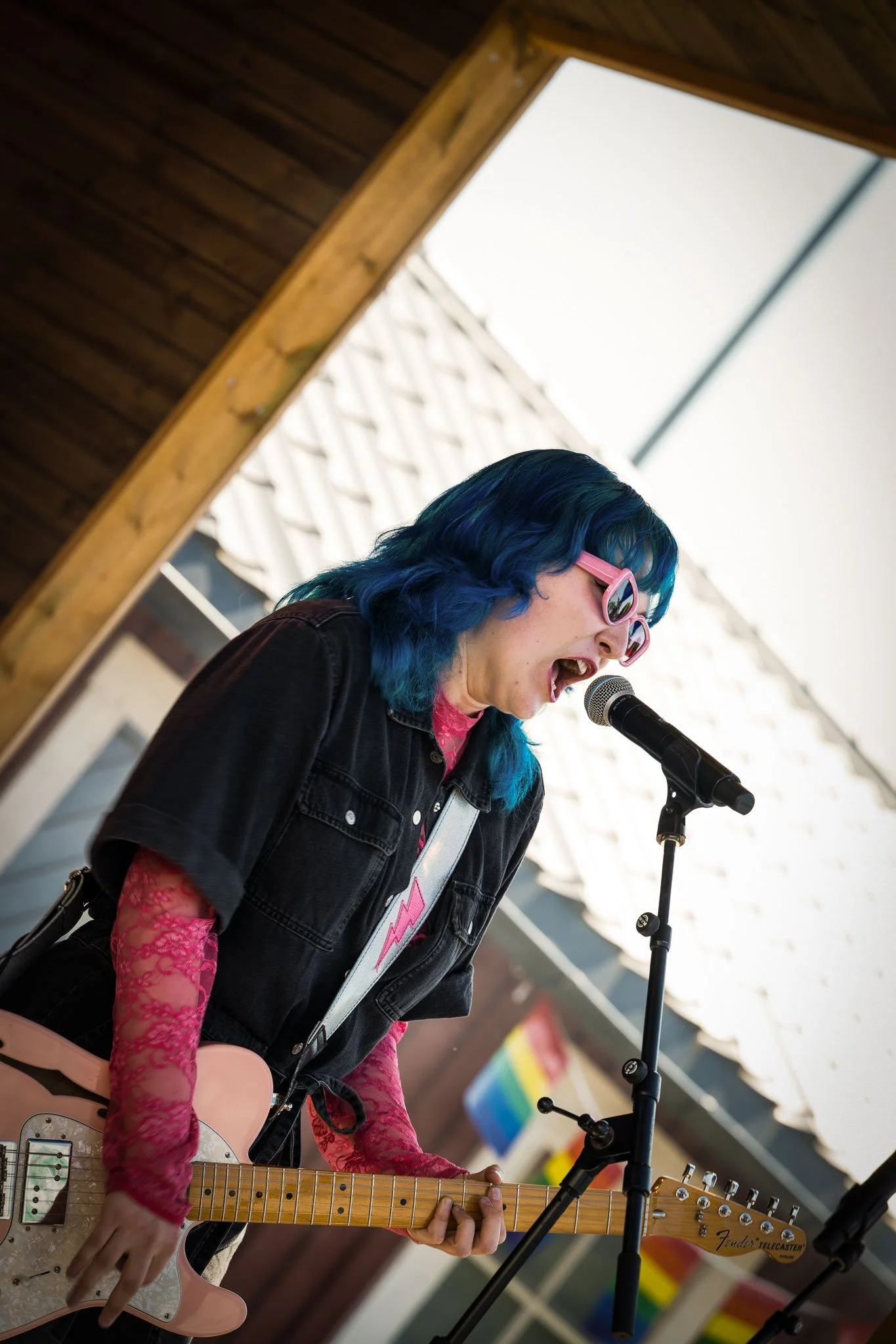 En person med blått hår, rosa solbriller og svart denimjakke, spiller og synger inn i en mikrofon med elektrisk gitar på en utendørs scene.