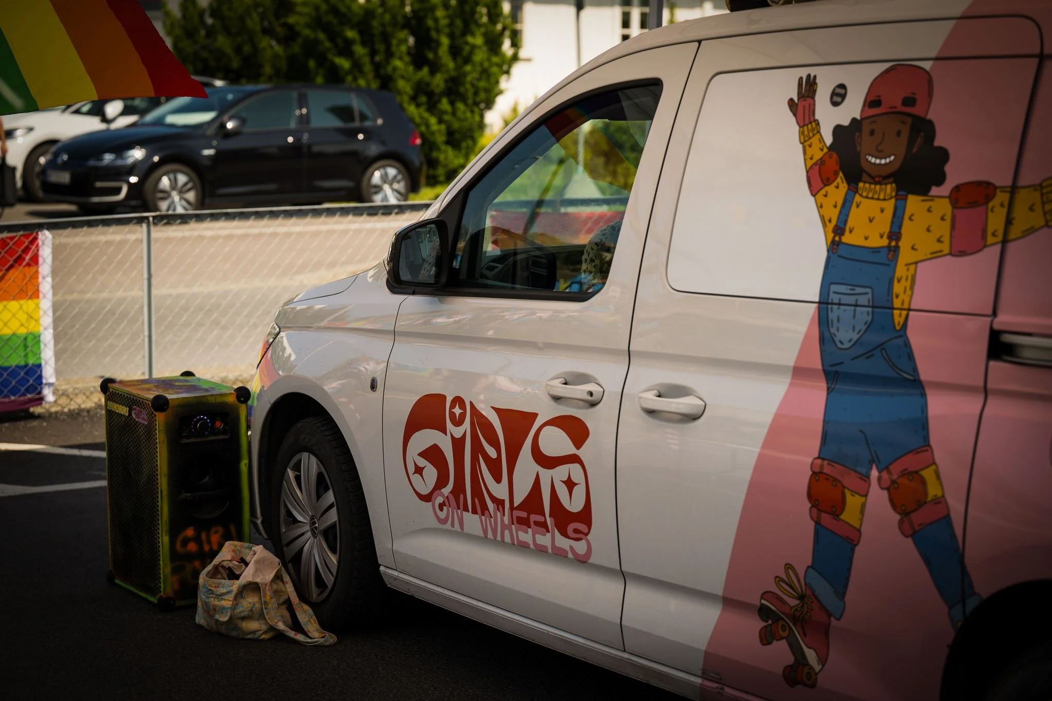 En varebil med grafikk av en jente på roller skøyter på siden, med teksten 'GIRLS ON WHEELS'. Parkert ved en utendørs scene med høyttaler og bagasje i nærheten. Noen andre biler kan sees i bakgrunnen.