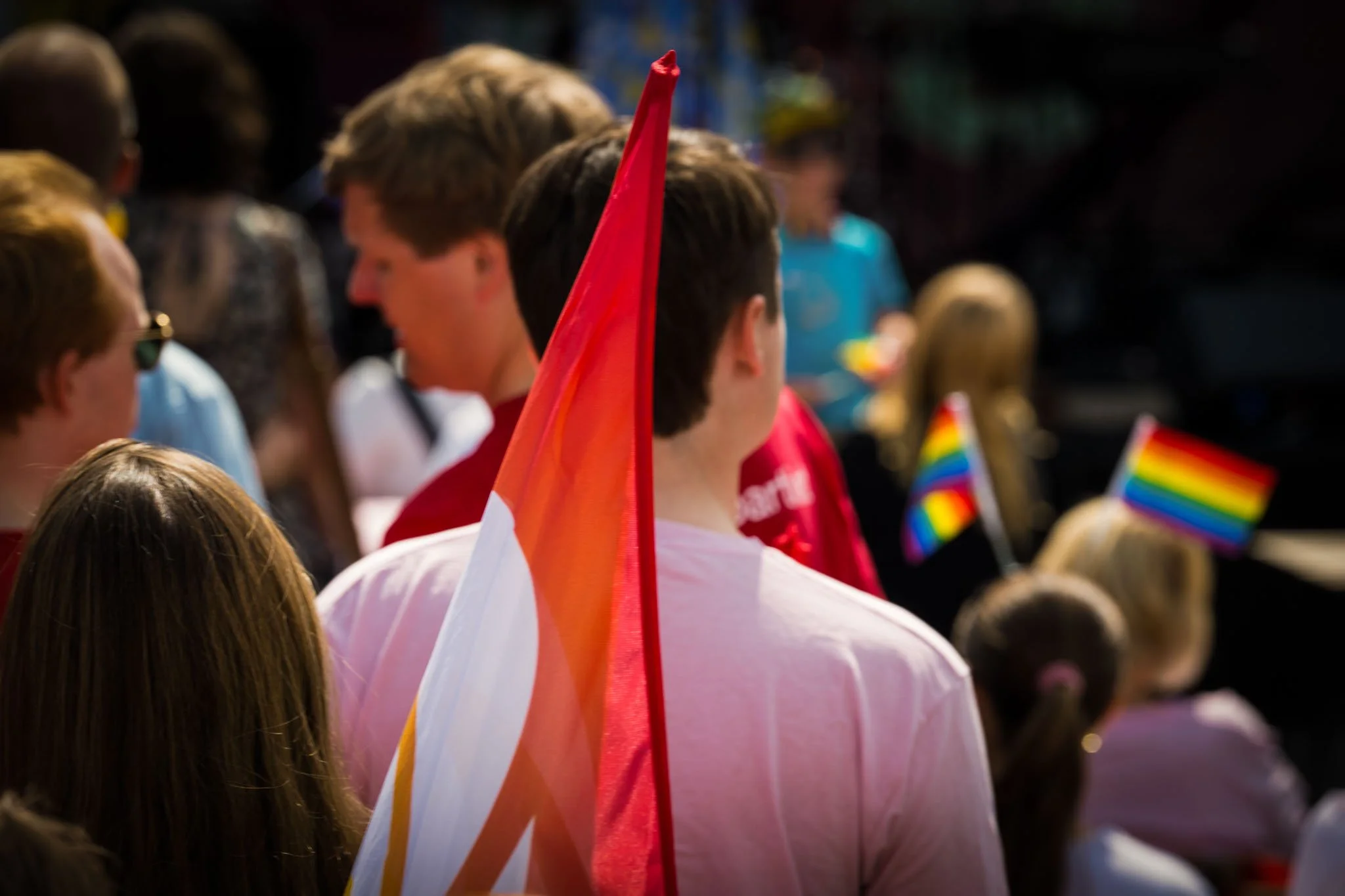 Personer på en parade eller samling, noen holder rainbow-fargede flagg, og det er et stort flagg i rødt, hvitt og oransje.