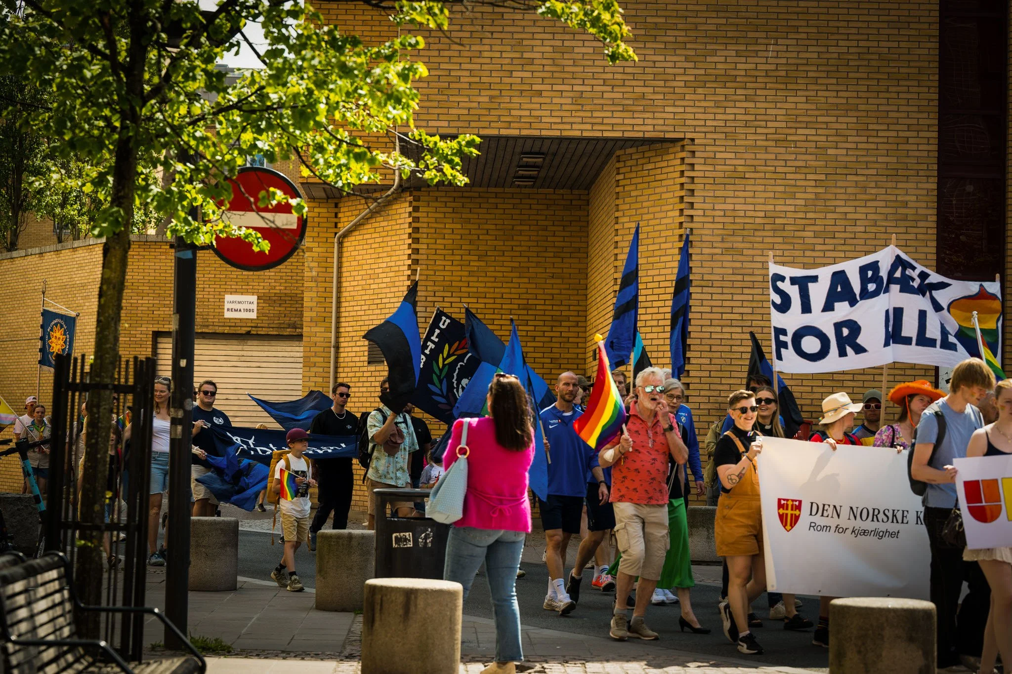 Personer delta i en demonstrasjon eller parade med flagg og bannere. Mange smilende mennesker, noen med regnbueflagg og andre med blå og svarte flagg, går langs en gate med gule mursteinbygninger i bakgrunnen. Noen mennesker tar bilder.