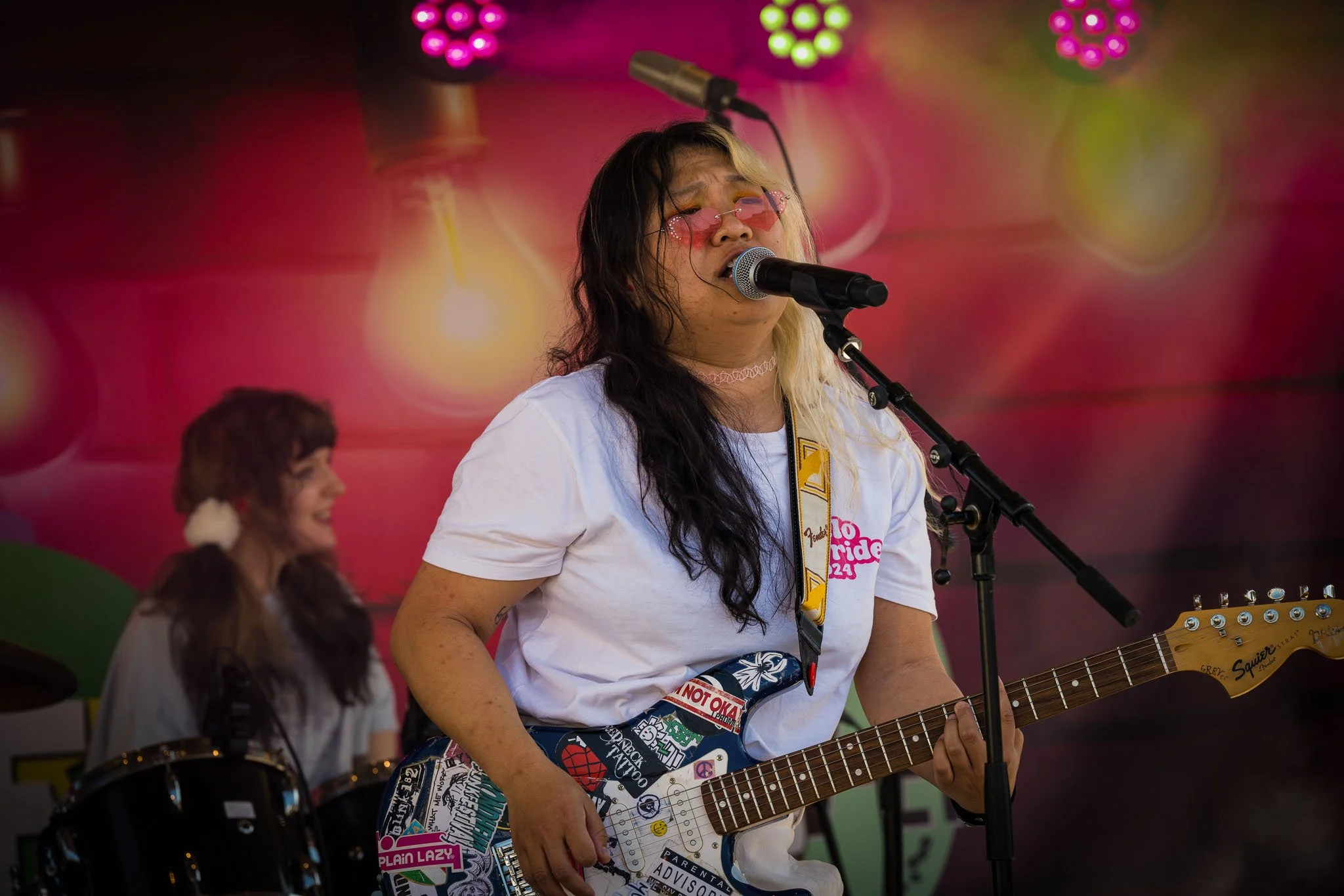 En person spiller gitar og synger inn i en mikrofon på en scene. Personens ansikt har rosa briller og lang, to-tonet hår, med en person i bakgrunnen som spiller trommer.