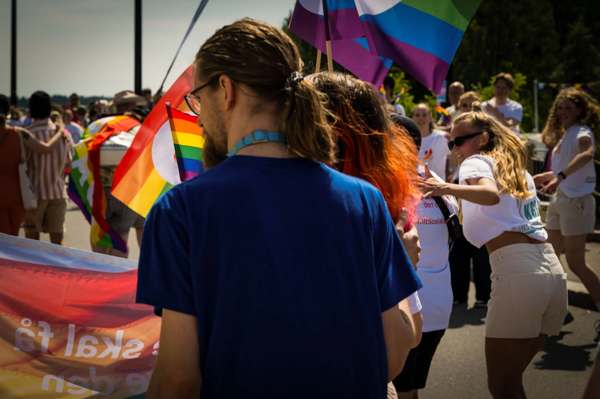 Ung dom og kvinner deltar i en LGBTQ+ rettighetsparade, holder regnbueflagg og smiler under solen.