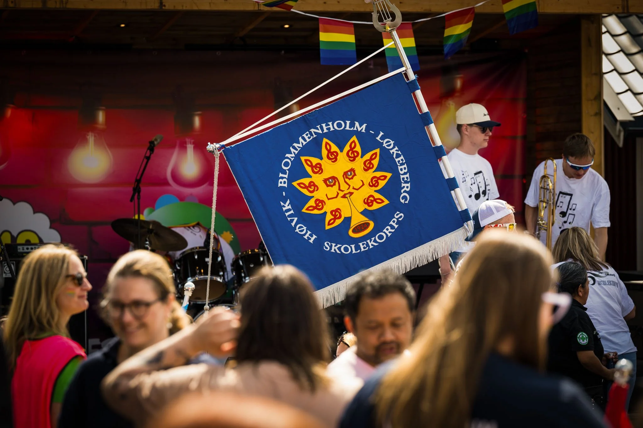 En gruppe mennesker deltar i en utendørs feiring med en blå flaggbanner som har teksten 'Høvik – Blommenholm – Løkkeberg Skolekorps' og en symbolsk sol i midten. De er kledd i hvite t-skjorter og solbriller, og mange av dem har på seg rainbow-element