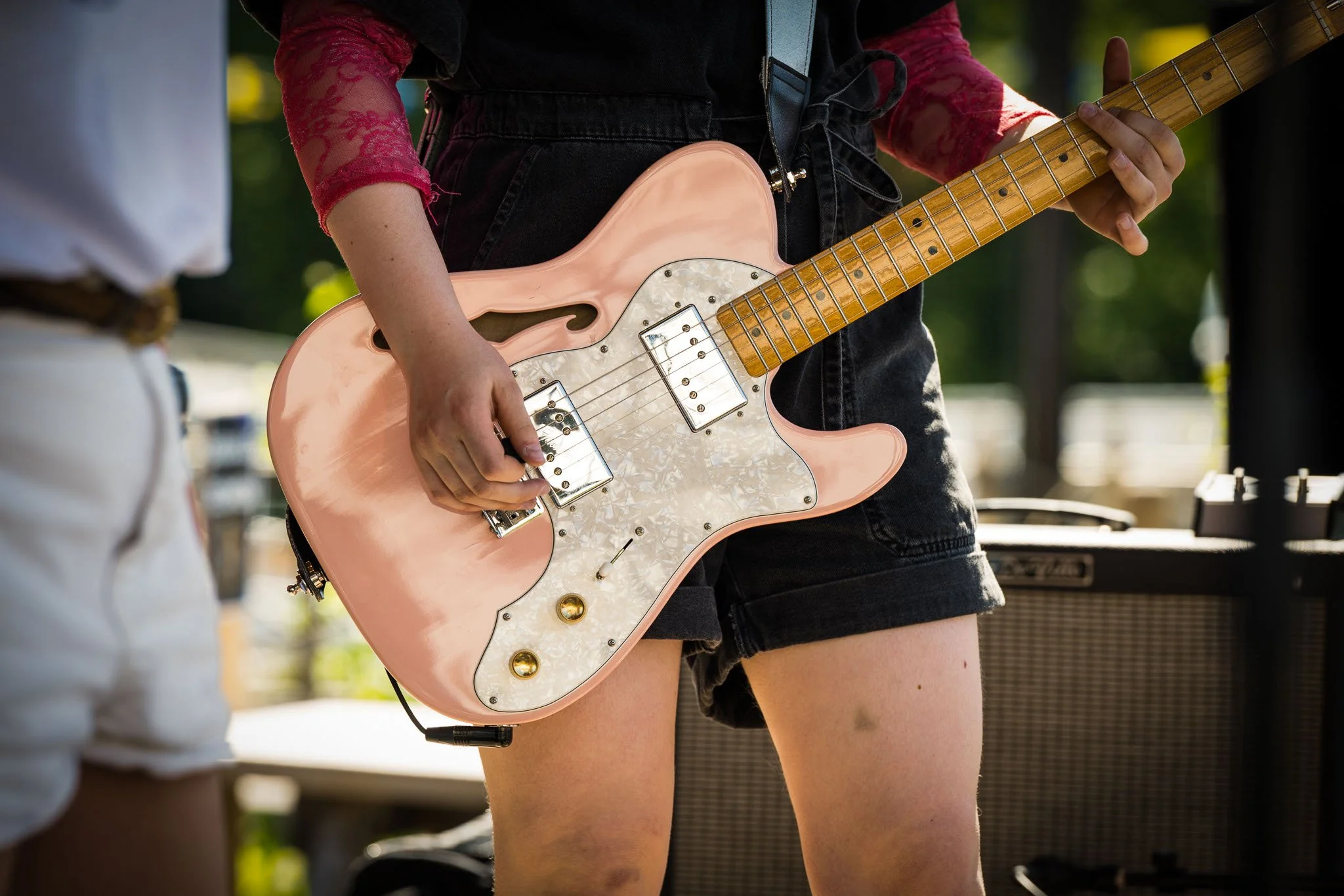 Person som spiller på en rosa elektrisk gitar, iført svart shorts og rød bluse med åpen erm.
