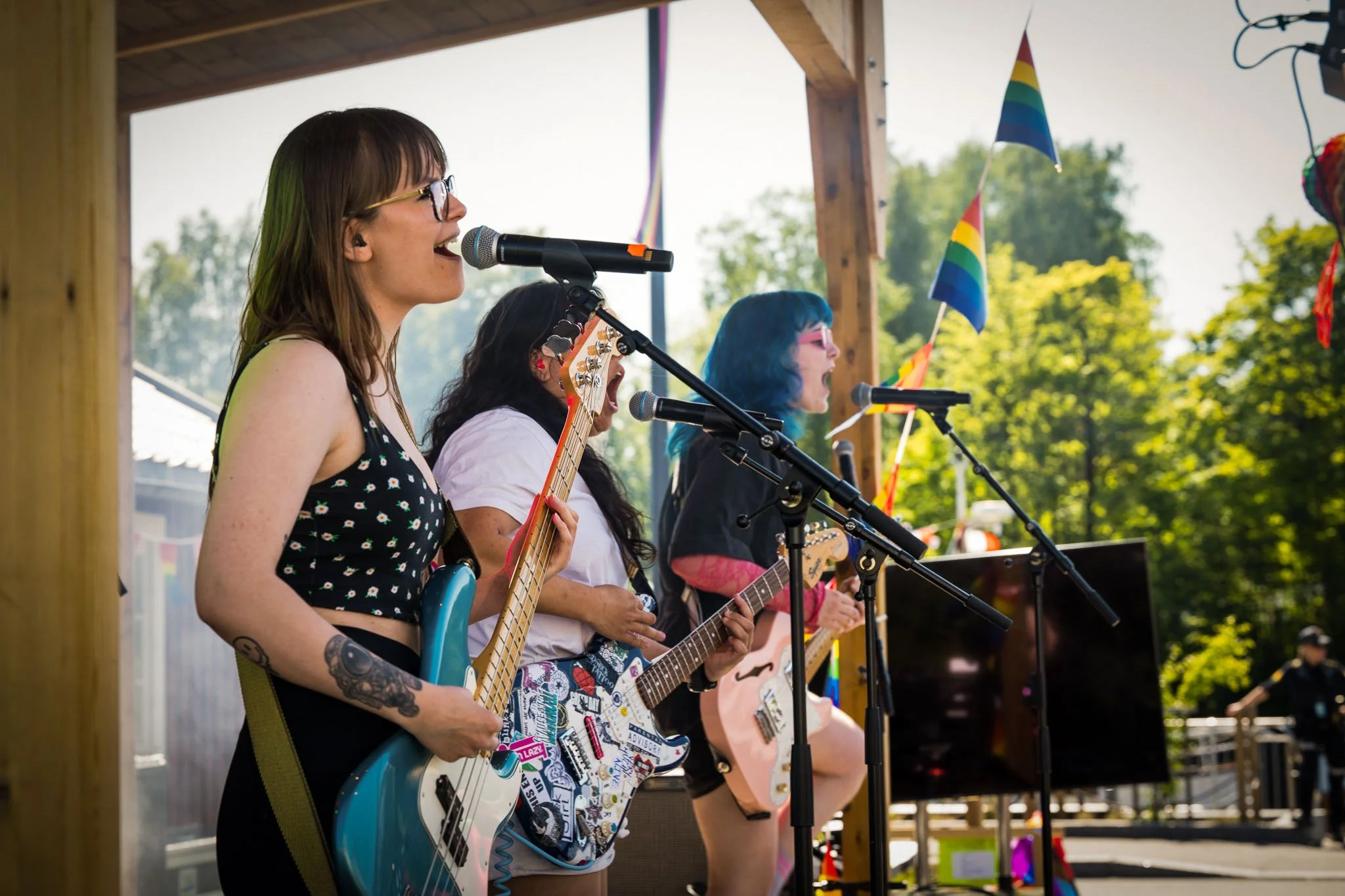 Tre kvinner som spiller elektriske gitarer og synger på en utendørs scene ved en festival, pyntet med regnbueflagger.