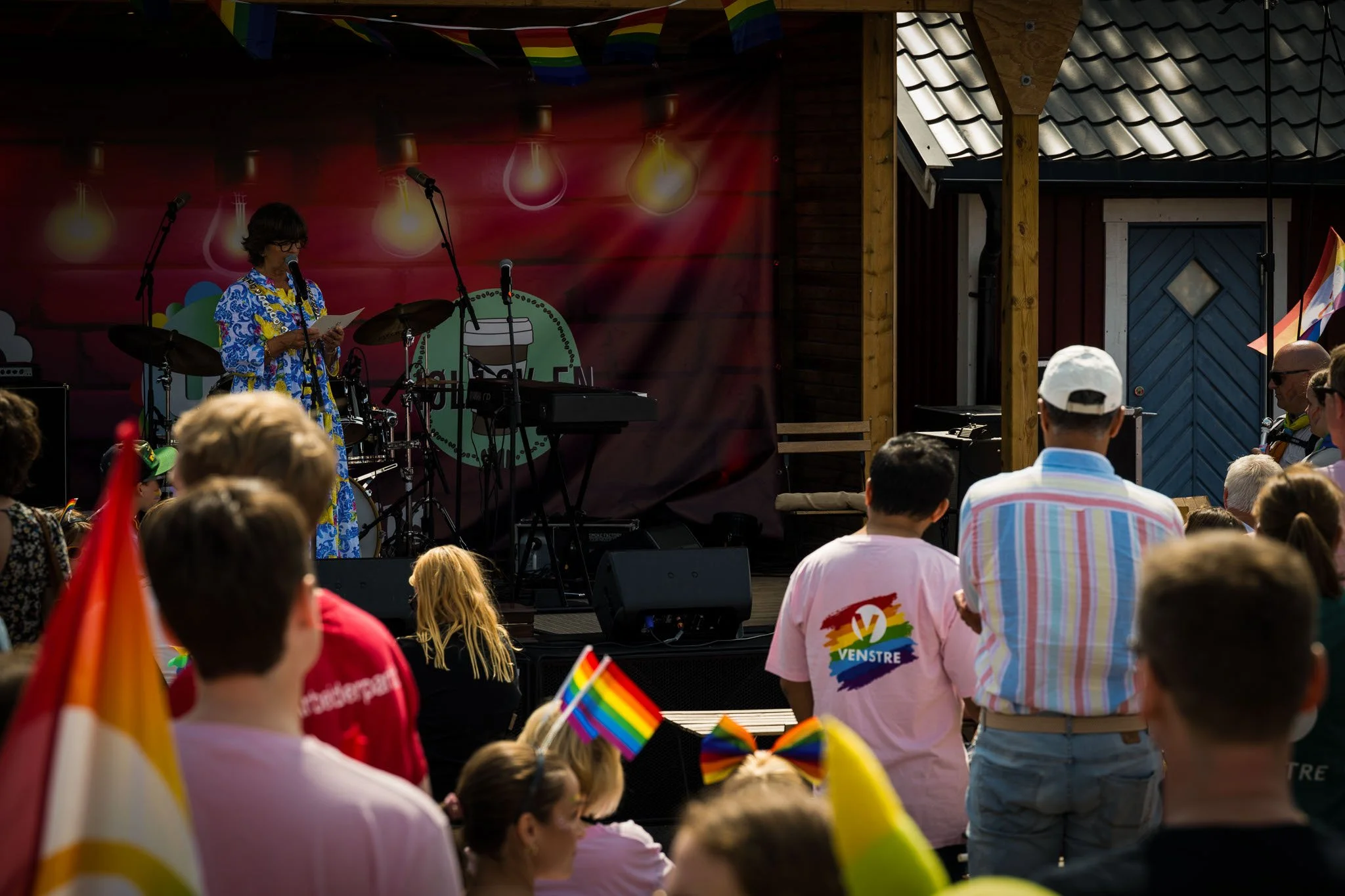 En utendørs scene under en Pride-feiring med en kvinnelig singer på scenen og folk i publikum som holder Pride-flagg og dekorasjoner.