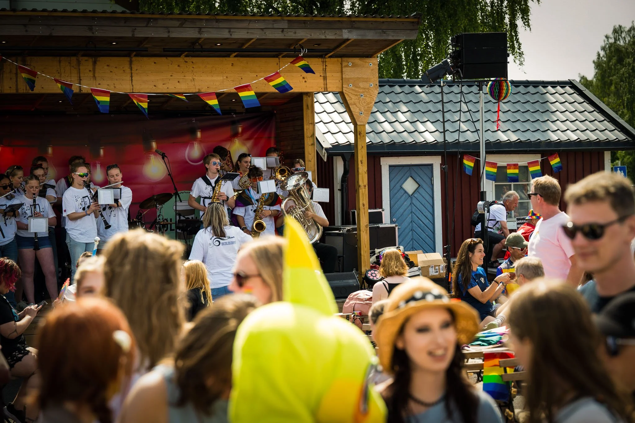Et utendørsarrangement med en musikalsk opptreden av en stor gruppe musikere på en scene, dekorert med prismefargede flagg, under en solrik dag med mange folk i aktivitet.