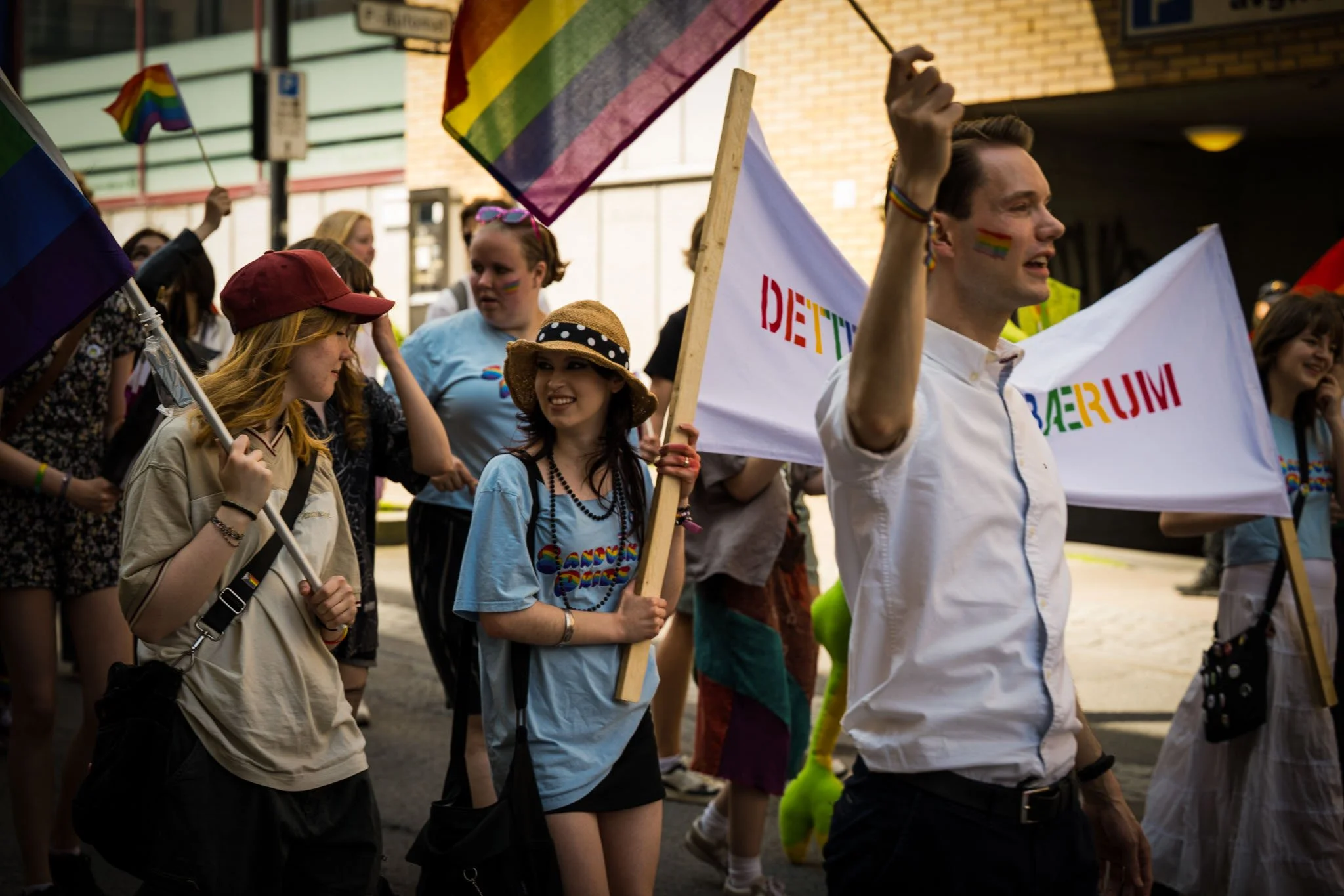 Grupper av mennesker deltar i pride-parade med regnbueflagg og fargerike klær på en bygate.