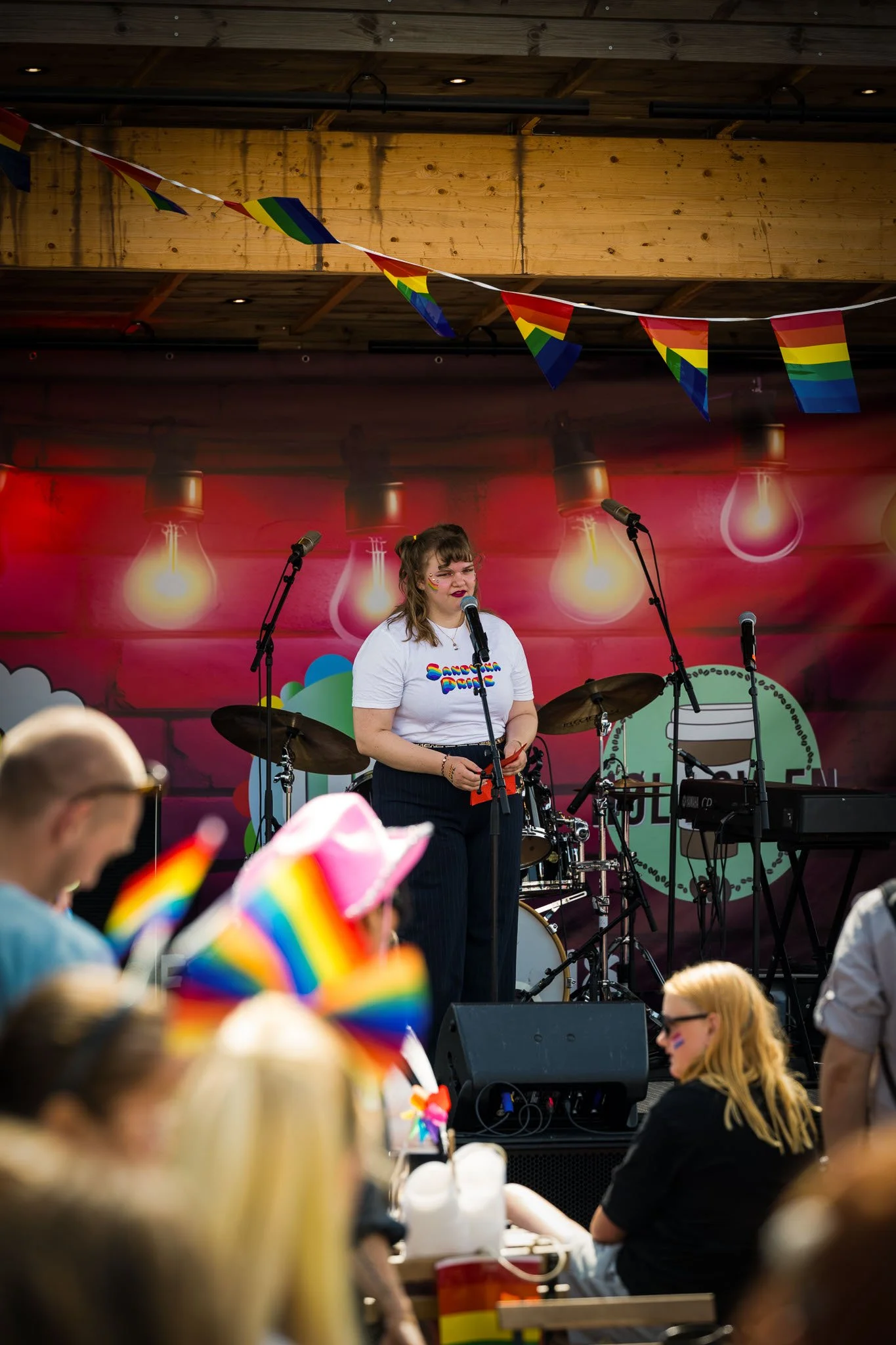 En kvinne står på en scene med mikrofon, omgitt av musikkutstyr, under en feiring med pride-dekorasjoner, inkludert regnbueflagg og flagg. Publikum ser ut til å feire pride, og det henger regnbuefargede flagg og dekorasjoner i bakgrunnen.