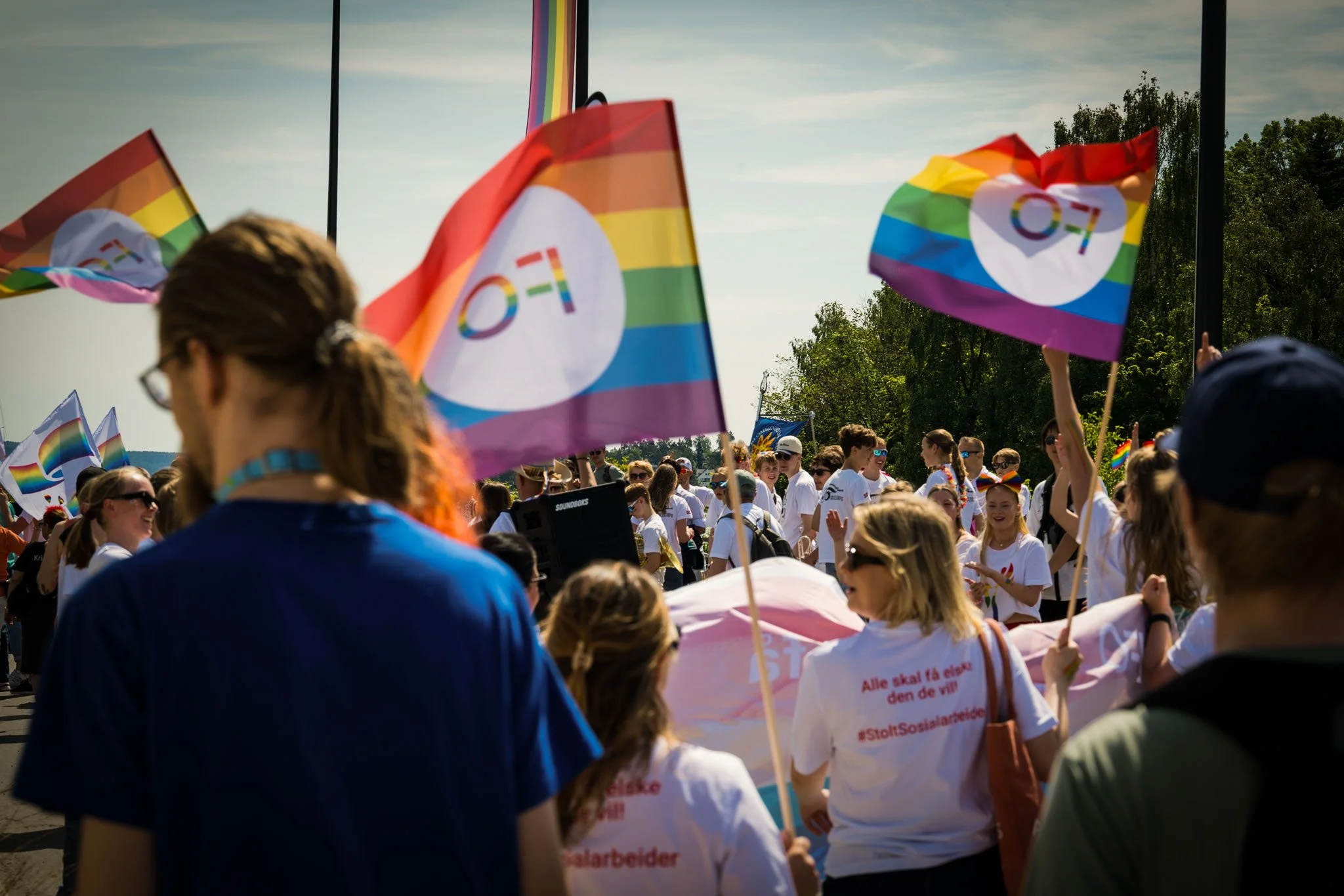 En gruppe mennesker deltar i en fargesterk parade eller demonstrasjon, mange bærer Pride-flagg og hvite t-skjorter med tekst. Deltakerne smiler og heier, mens solen skinner over en parkert plass med trær i bakgrunnen.