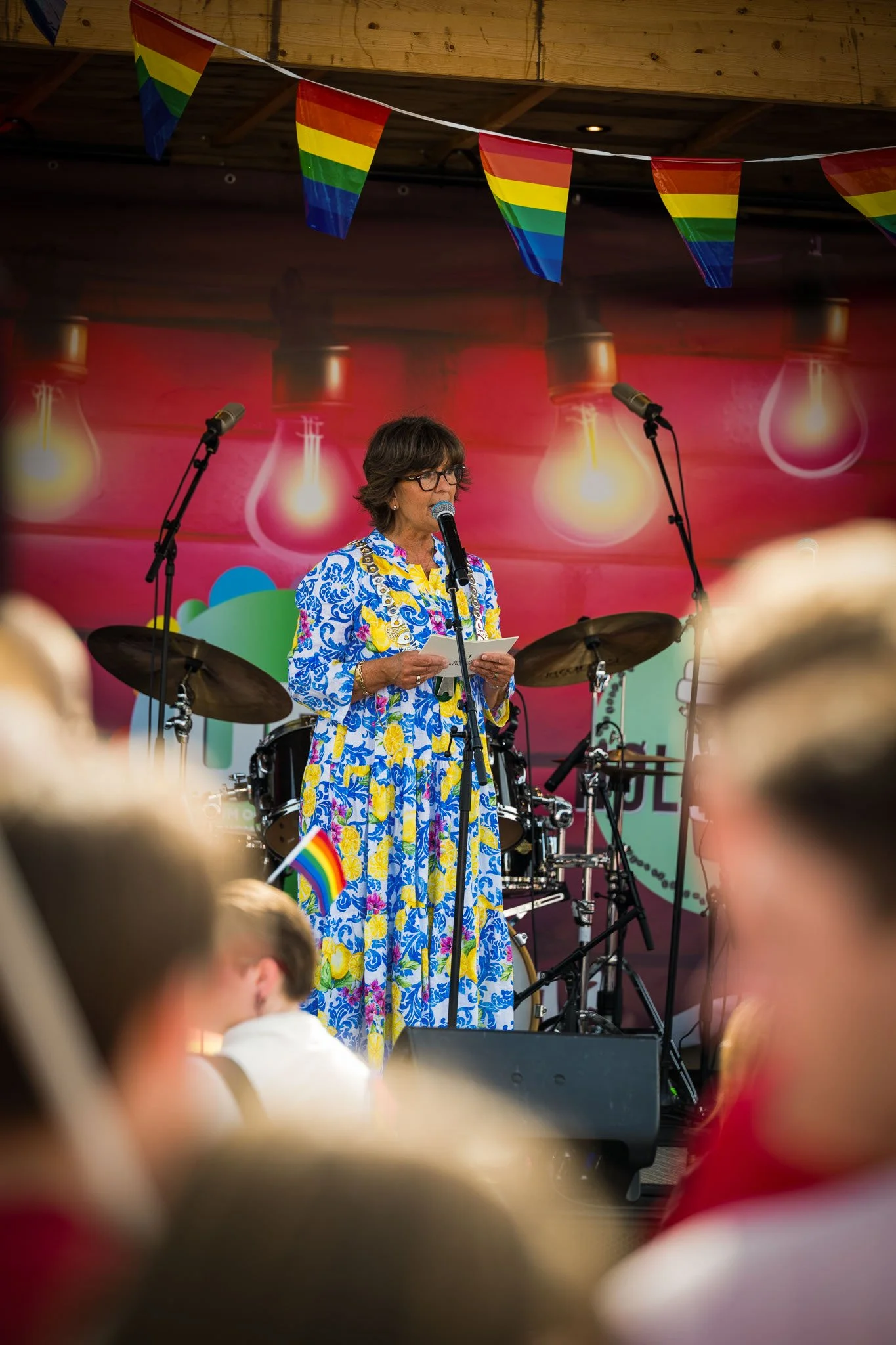 En kvinne står på en scene med mikrofon, omgitt av musikkinstrumenter, under en pride-feiring. Hun har på seg en fargerik, blomstrete kjole, og det henger pride-flagg over scenen.