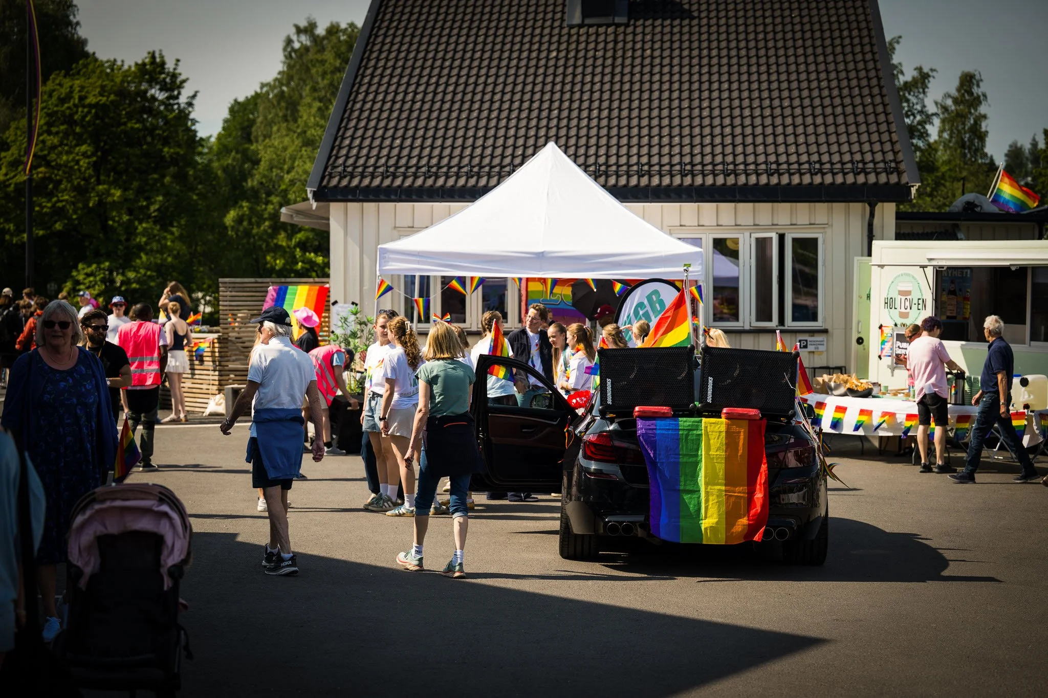 En gruppe mennesker deltar i en Pride-parade eller feiring, med mange regnbueflagger og pynt, i et utendørs område foran et hvitt hus med svart tak.