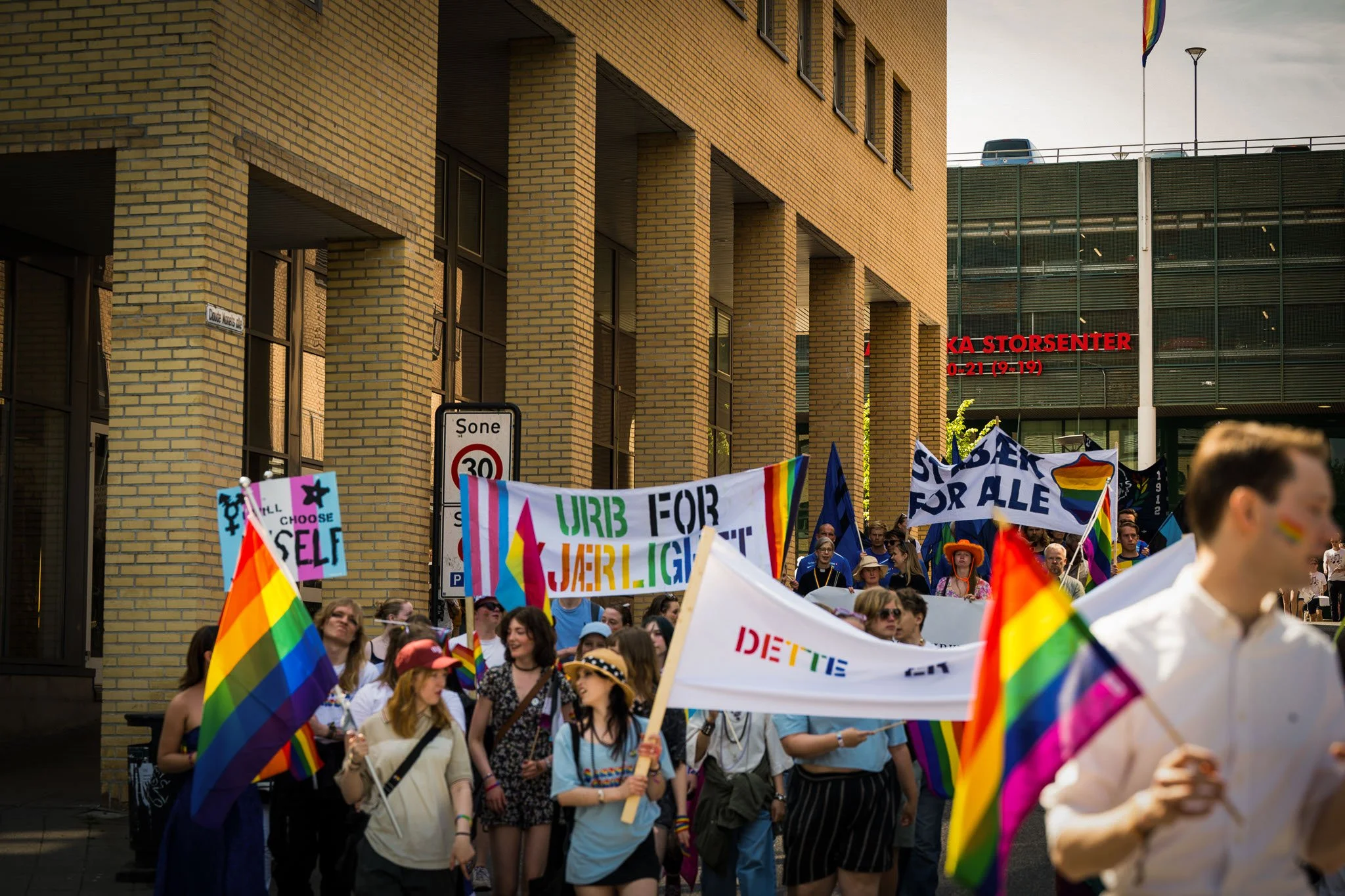 En gruppe mennesker deltar i en parade med regnbuefargede flag og banner for LGBTQ+ rettigheter, på en gate med bygninger i bakgrunnen.