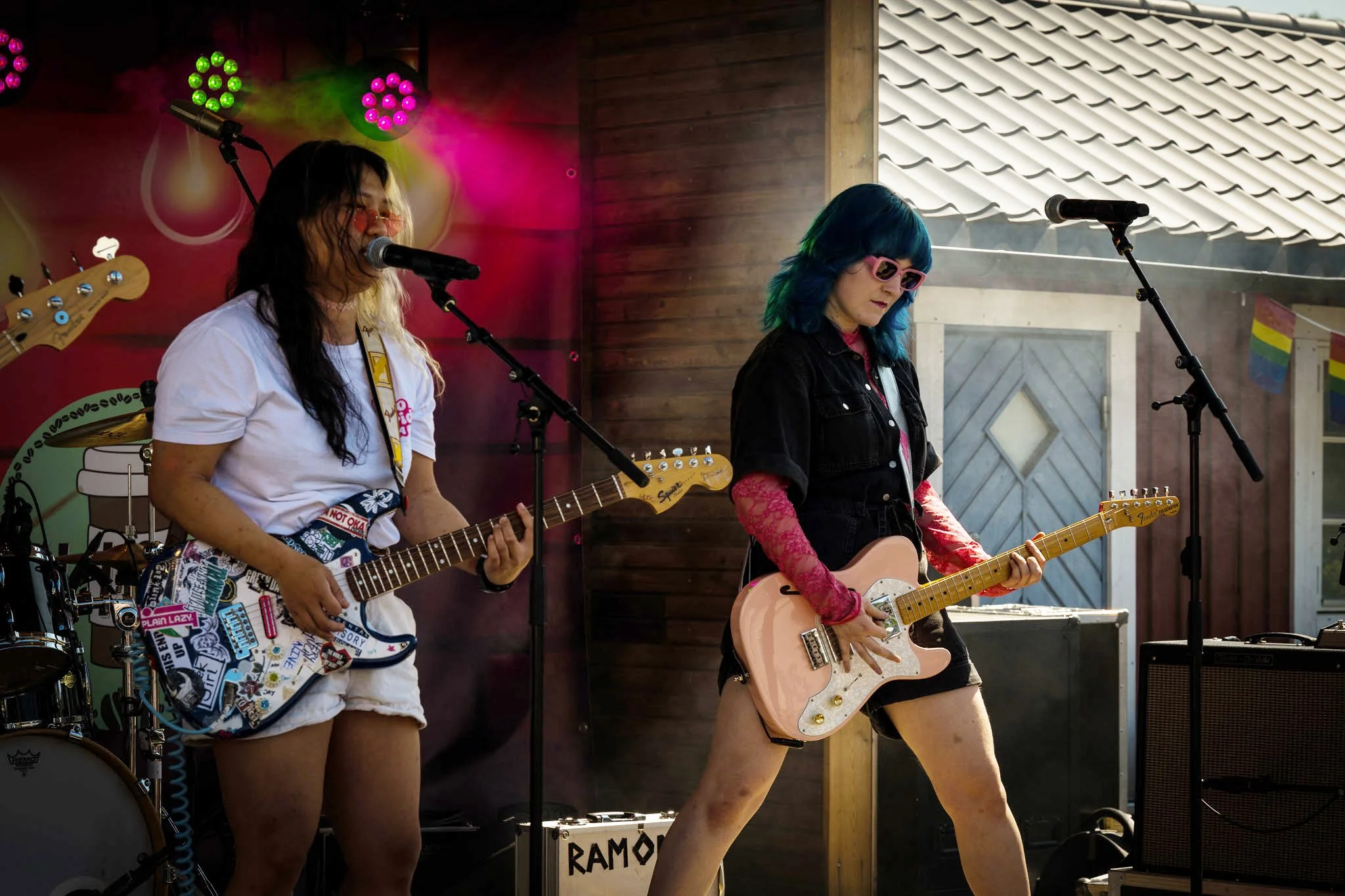 To kvinner som spiller elgitarer på en scene. Den ene har langt mørkt hår og en hvit t-skjorte, den andre har blått hår og rosa solbriller. Begge ser ut til å ha det gøy mens de spiller.