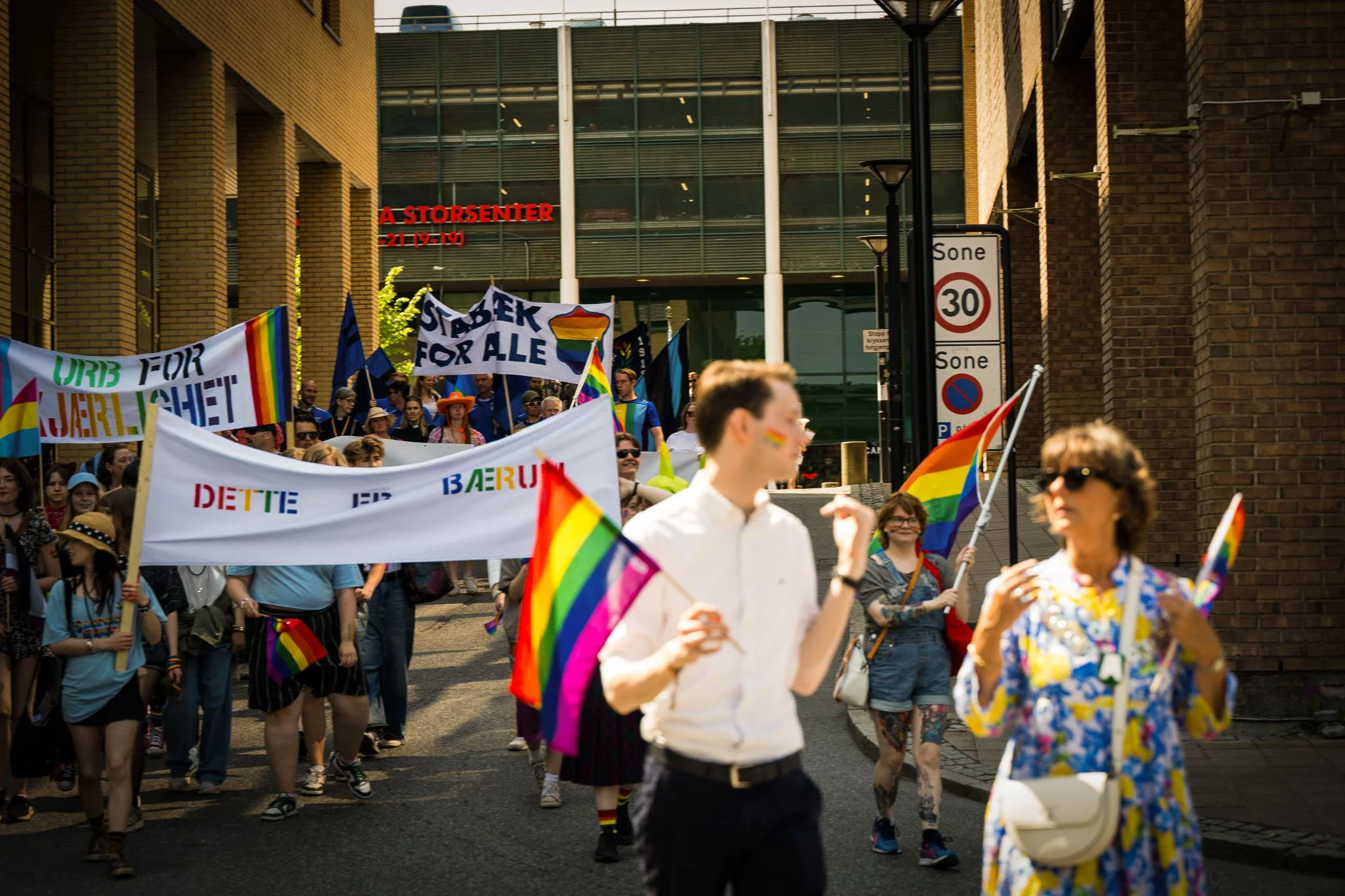 Personer som deltar i en parade med LGBTQ+ flagg og regnbuefargede bannere, gående nedover en gate med bygninger i bakgrunnen.