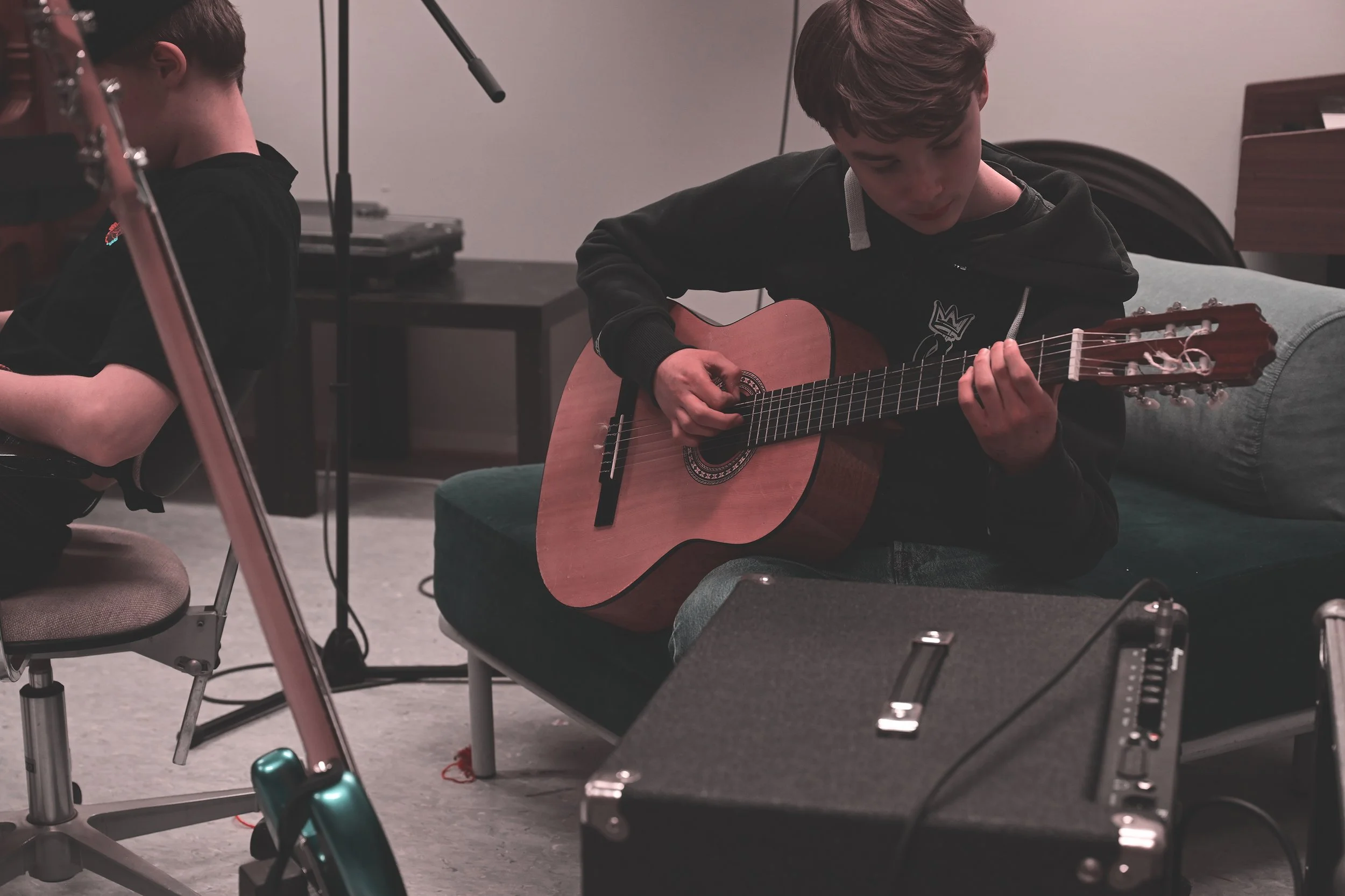 To ungdommer spiller gitar i et rom, med en forsterker foran dem, en i midten med akustisk gitar og en på venstre med elektrisk gitar. Rommet er utstyrt for musikkøving.