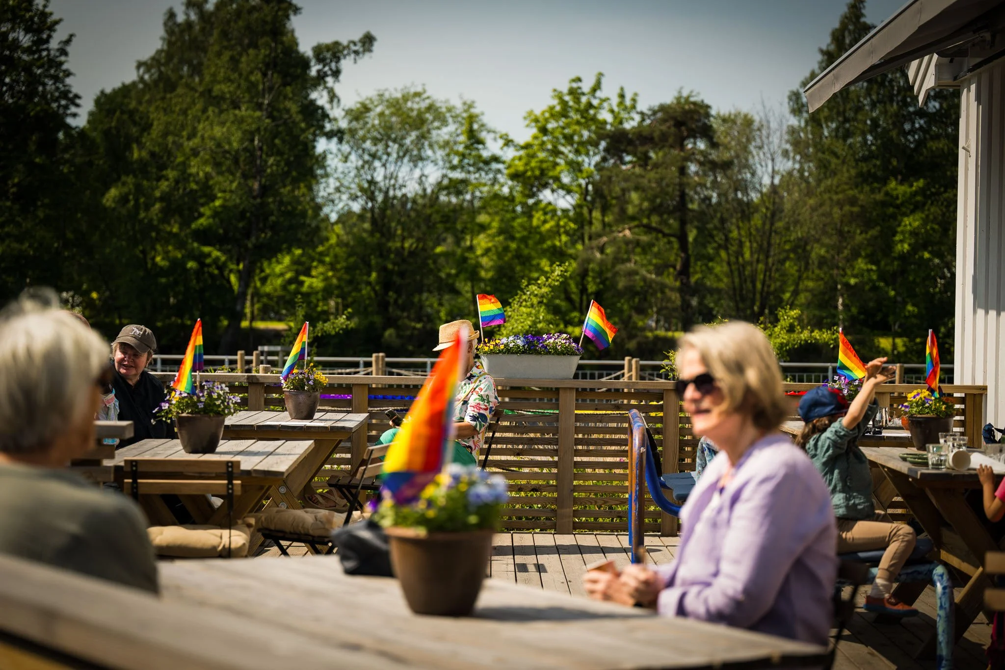 Ute på en terrasse med folk som feirer Pride, dekorert med små rainbow-flag i blomsterkasser og på bordene, omgitt av grønne trær.
