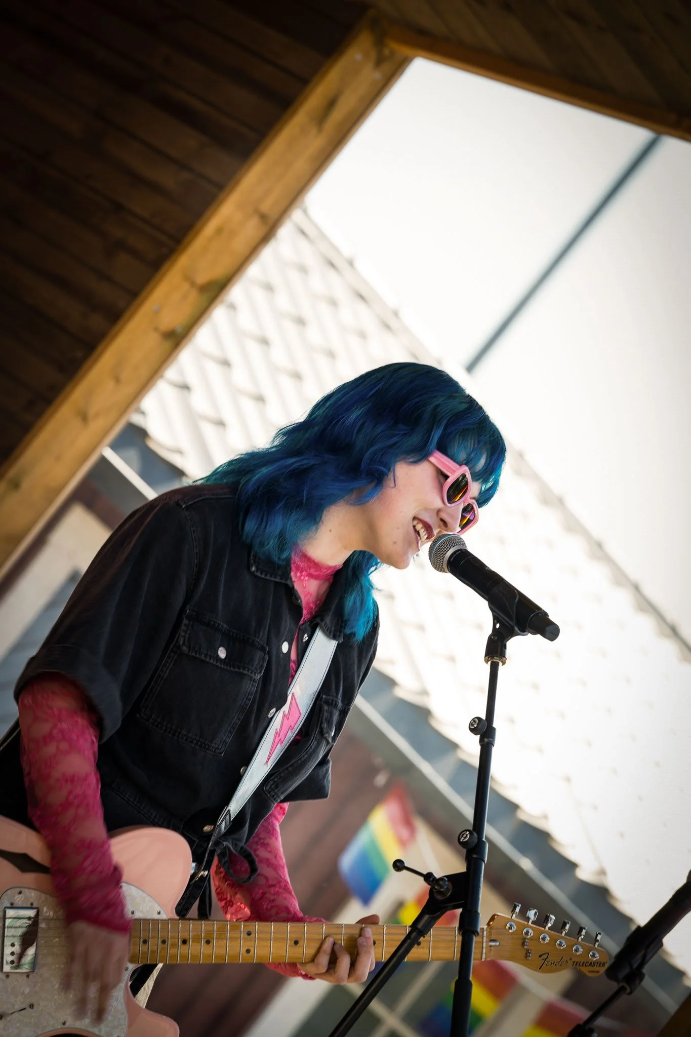 En kvinne med blått hår og rosa solbriller spiller gitar og synger inn i en mikrofon under en konsert.