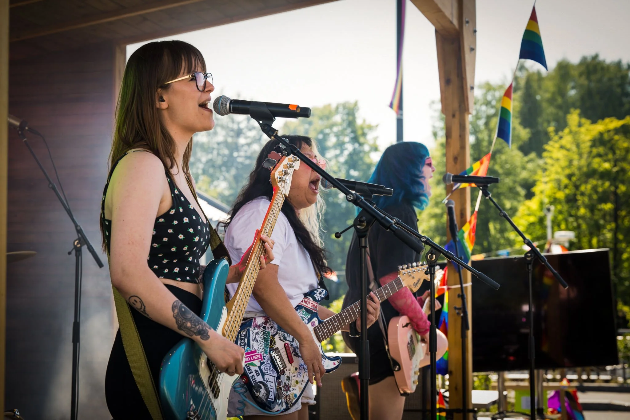 Tre kvinner som spiller gitar og synger på en utendørs scene under en pride-feiring, med pride-flagg i bakgrunnen.