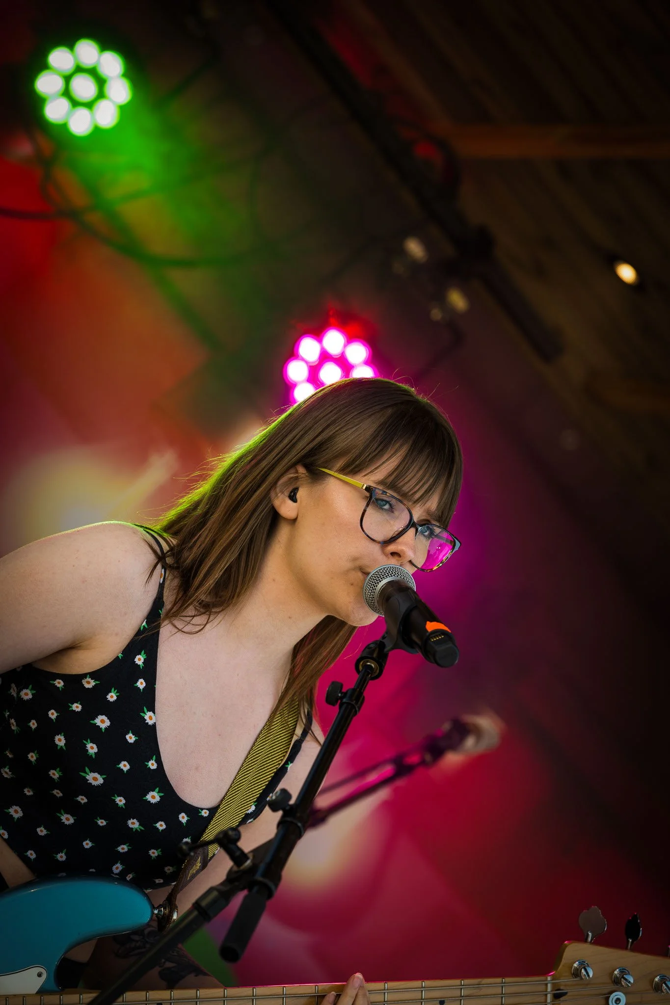 En kvinne med briller synger inn i mikrofon mens hun spiller gitar på scenen med fargede lys bak henne.
