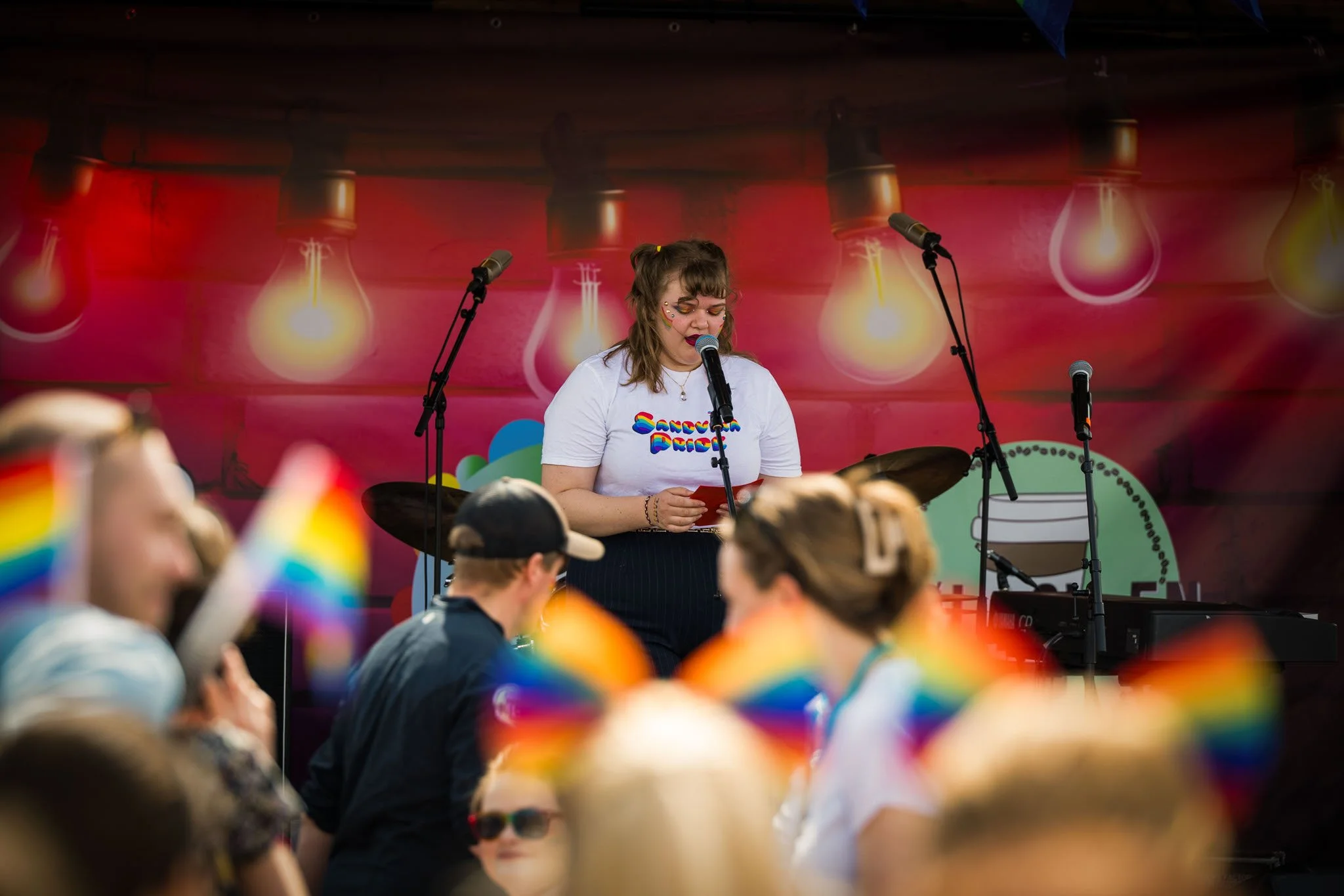 En kvinne synger foran et publikum under en Pride-feiring, med personer som bærer regnbuefargede hatter i forgrunnen.
