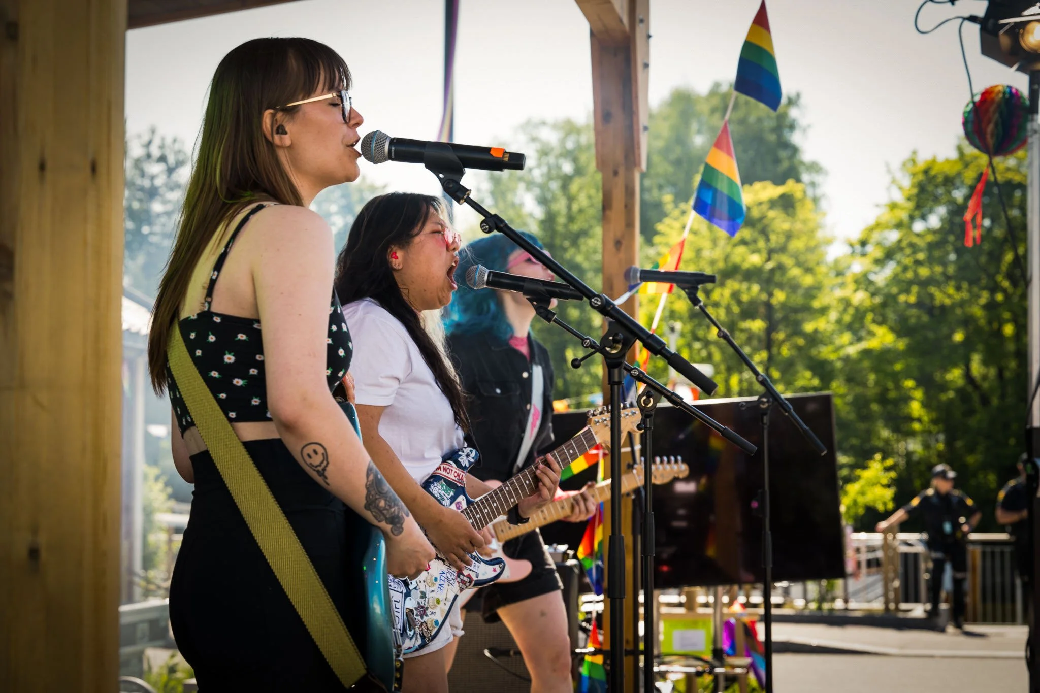 Tre kvinner spiller gitar og synger på en utendørskonsert, med Pride-flagger i bakgrunnen.