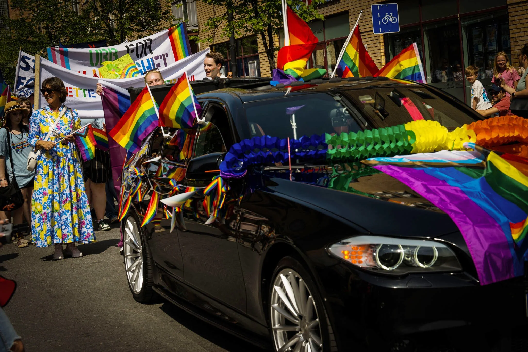 En svart bil dekorert med regnbuefargede flagg og pynt, omgitt av mennesker som holder regnbueflagg, deltar i en parade til støtte for LHBTQ+-rettigheter.