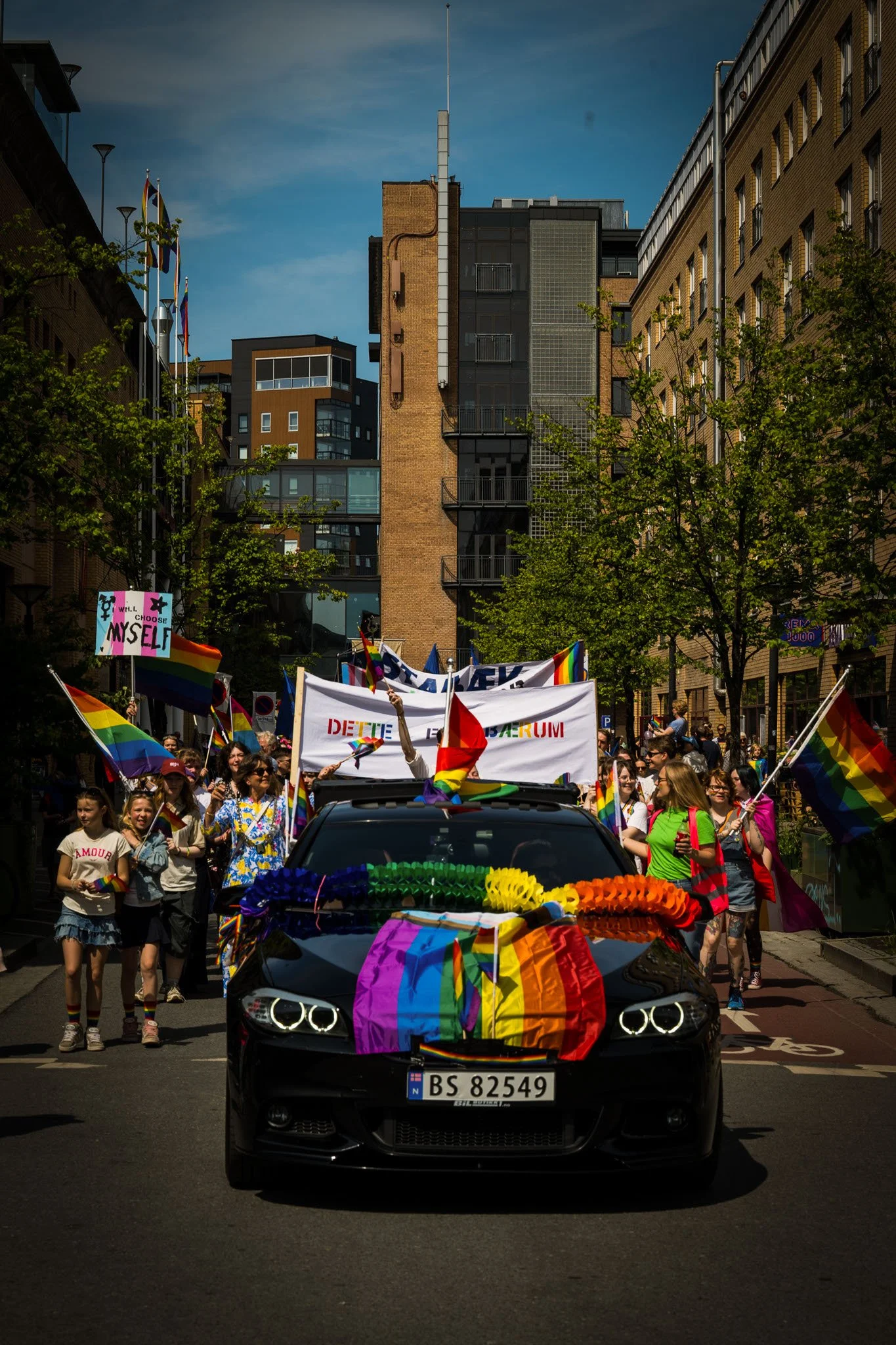 En parade med mennesker som holder opp regnbueflagg og bander under en farget bil med regnbuekrans i sentrum, på en gate omringet av bygninger.