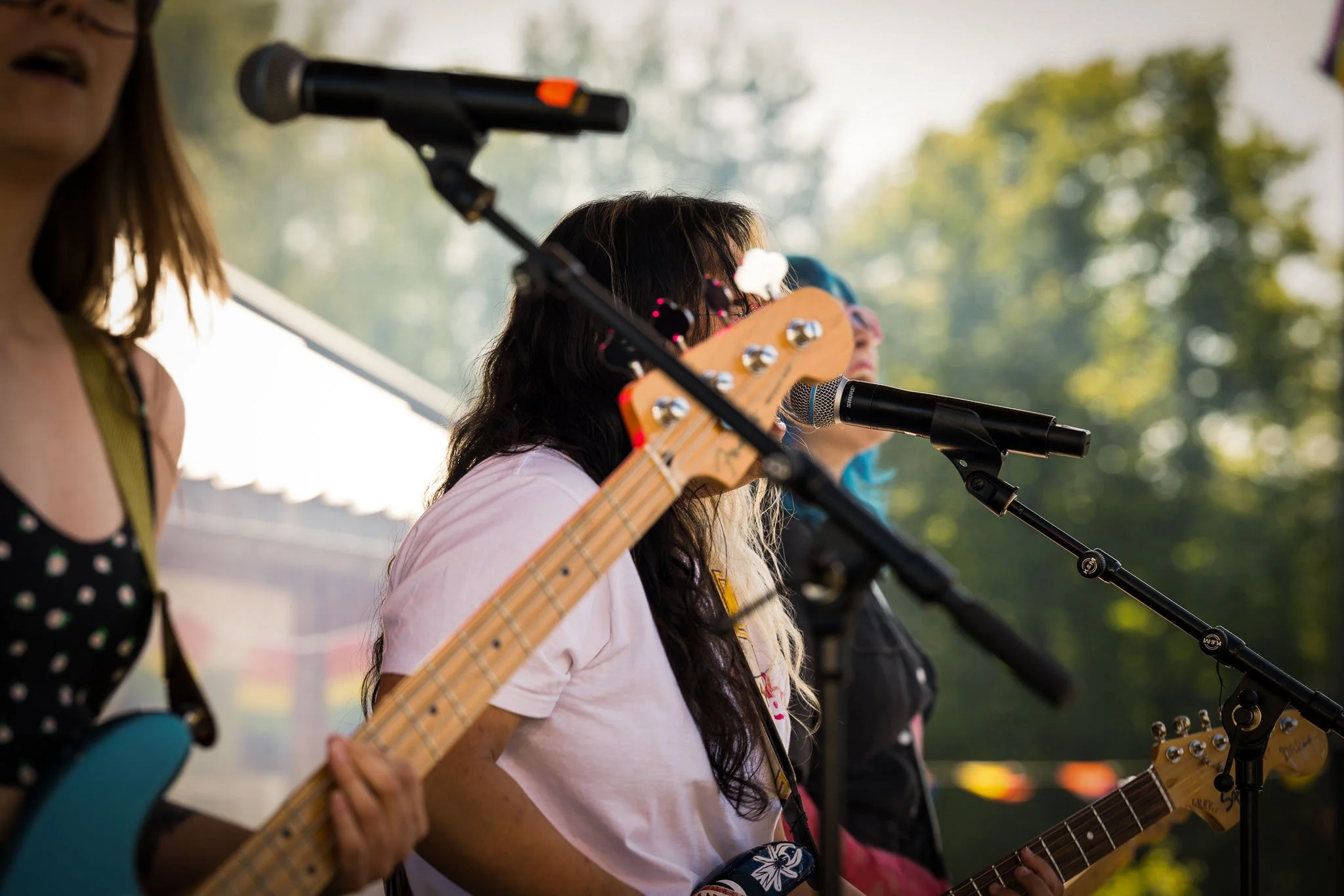 Tre kvinner spiller gitar og synger på en utendørs scene med greener i bakgrunnen.