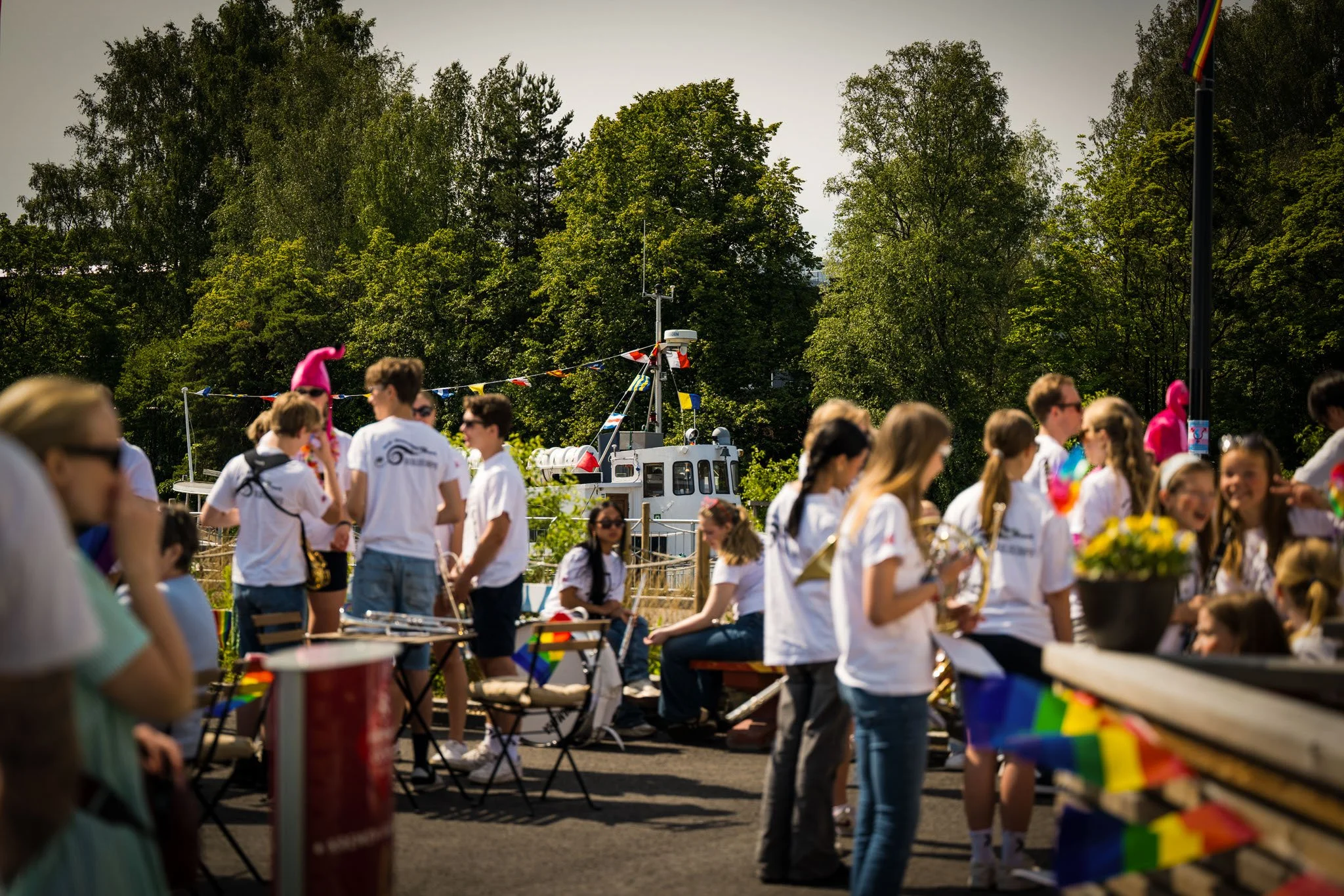Gruppe av mennesker i hvite t-skjorter samlet utendørs under en festival eller feiring, med trær og en bakgrunnsbåt.