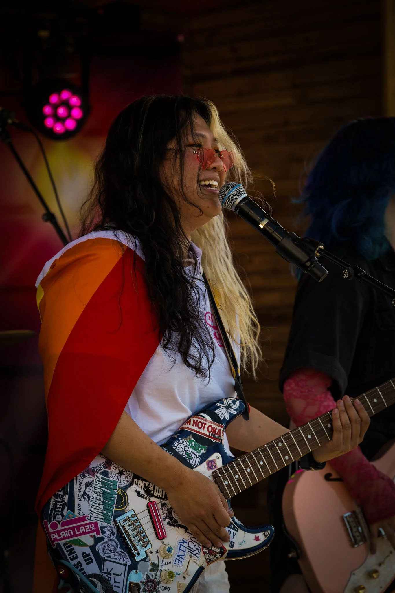 En person spiller elektrisk gitar med mange klistremerker, synger inn i mikrofonen, og har på seg rosa hjerteformede solbriller, foran en trevegged bakgrunn.
