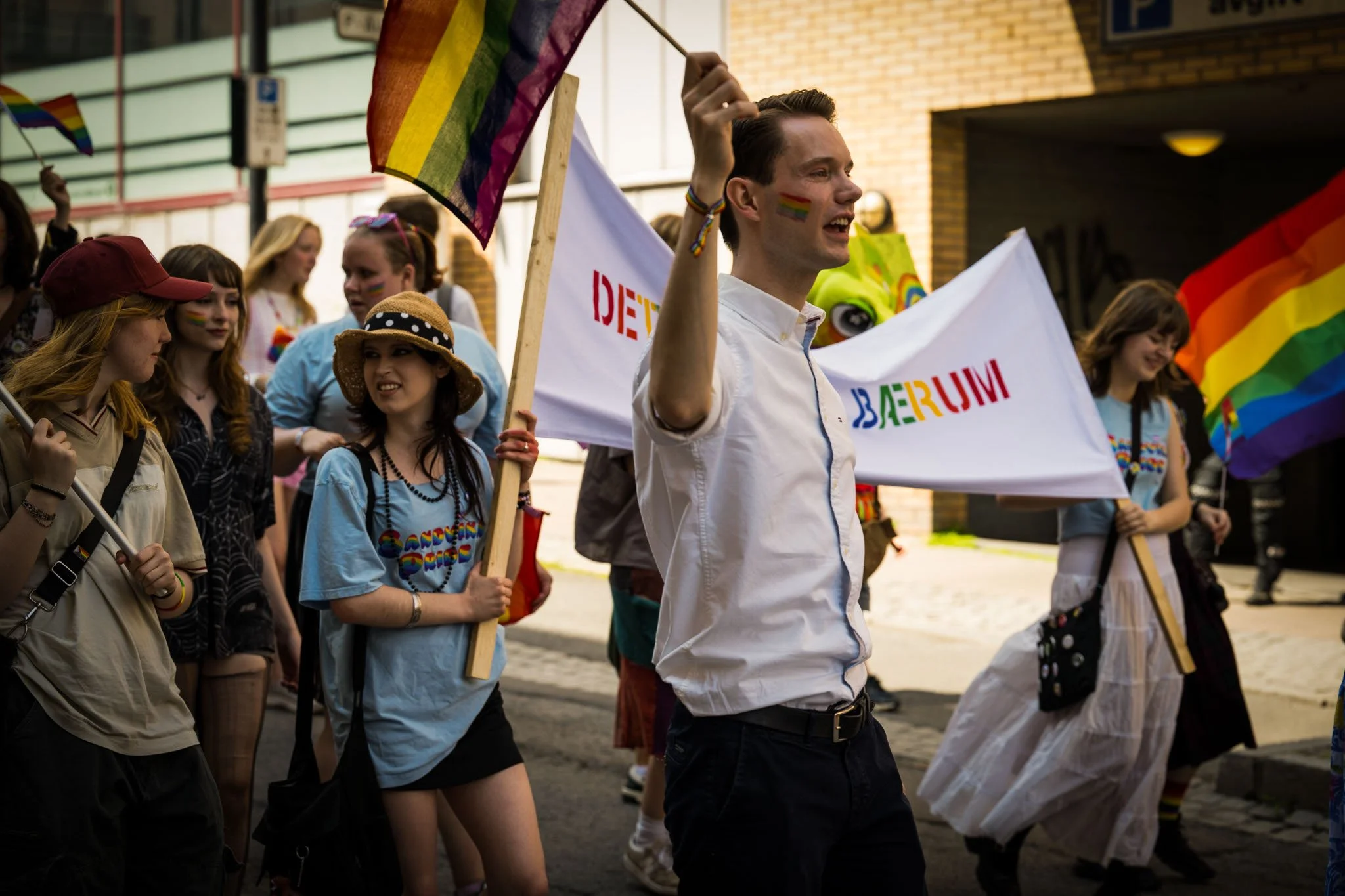Gruppemedlemmer i en parade med regnbueflagg, noen med regnbuefargede ansiktstegninger og klær, feirer kjærlighet og mangfold.