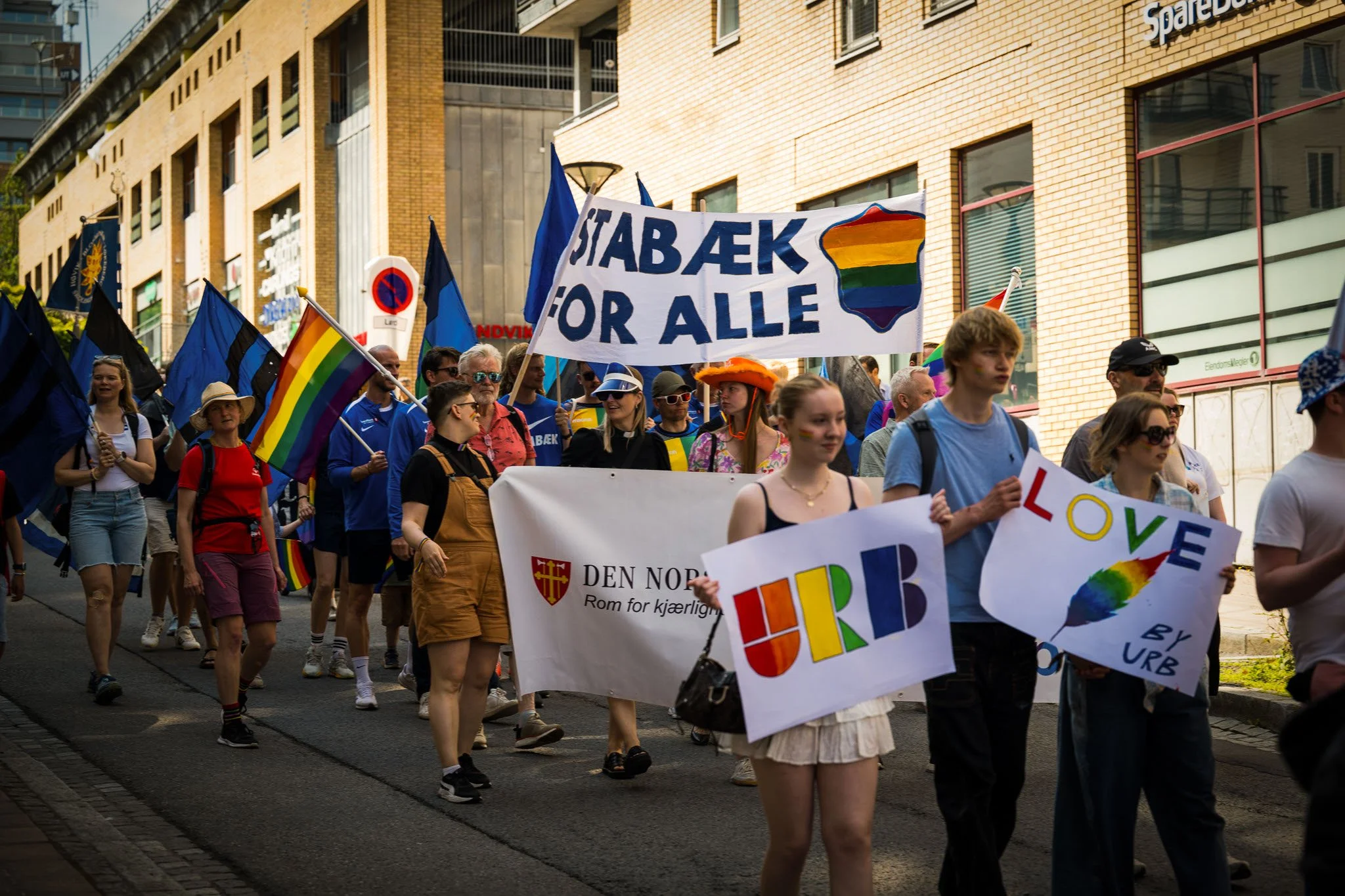 Folk i en parade som bærer regnbueflagg og banner med meldinger om kjærlighet og like rettigheter, deltar i en felles markering for LGBTQ+ rettigheter på en bygate.