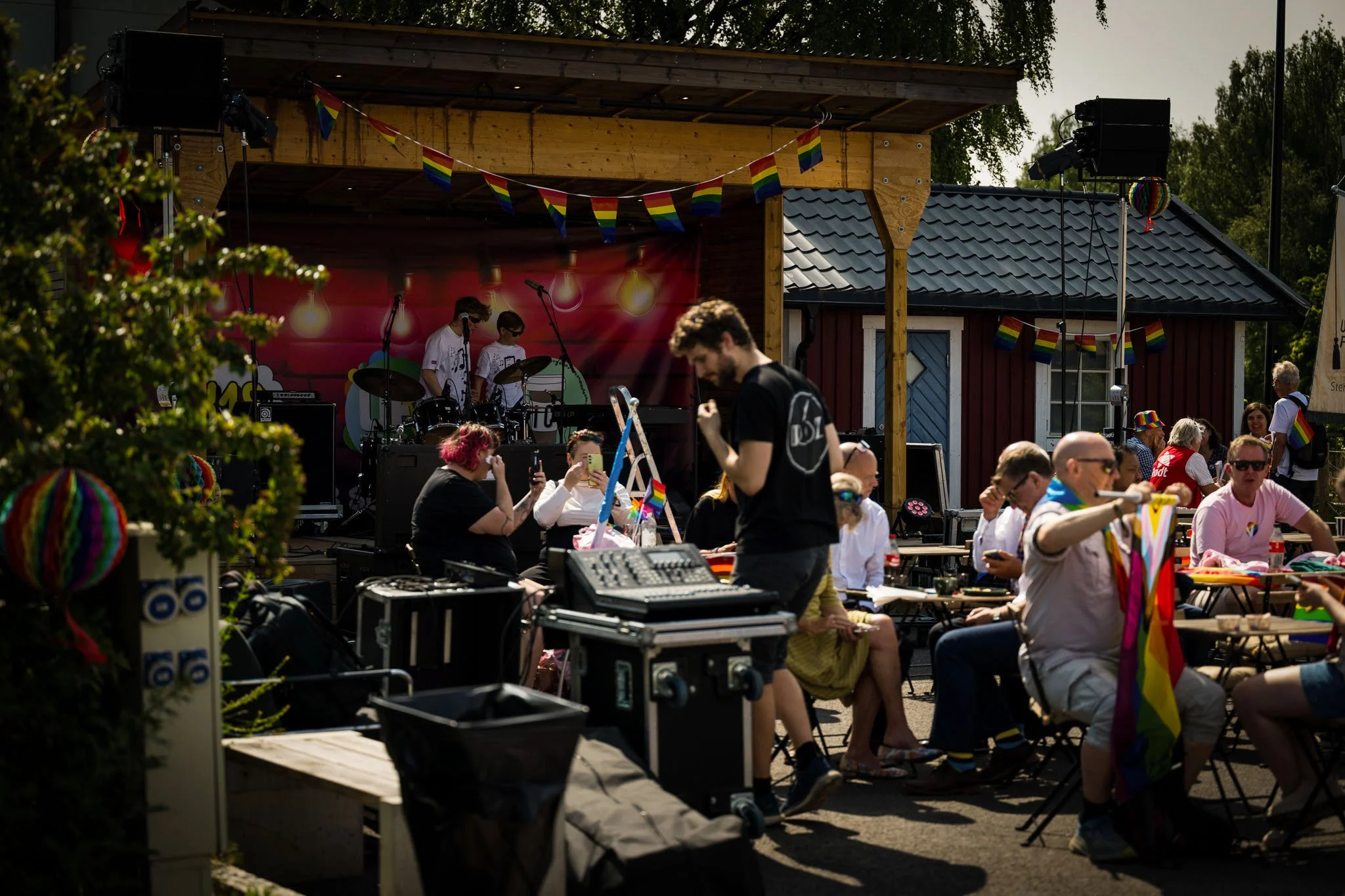 En utendørs scene med folk som samles og deltar i et LGBTQ+-arrangement. Det er en scene med et band som opptrer, og mange mennesker sitter ved bord. Gjestene bærer regnbuefargede flagg og pyntetøy, og det er pynt og bannere med regnbuefargede flagg.