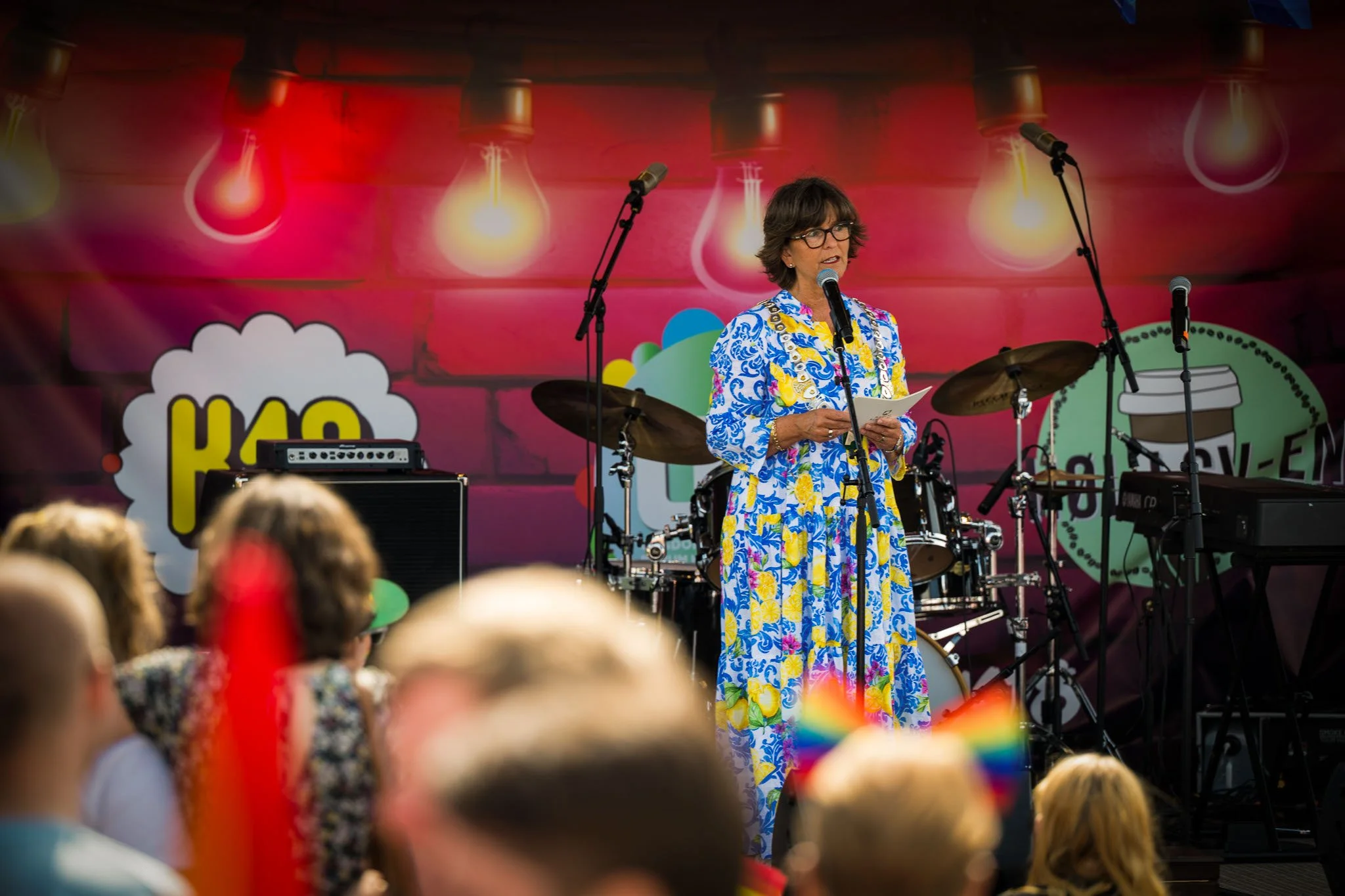 En kvinne står på en scene med mikrofon, omgitt av musikkinstrumenter, under en utendørs forestilling. Bakgrunnen har fargerike lys og grafiske elementer, og publikum ser på.