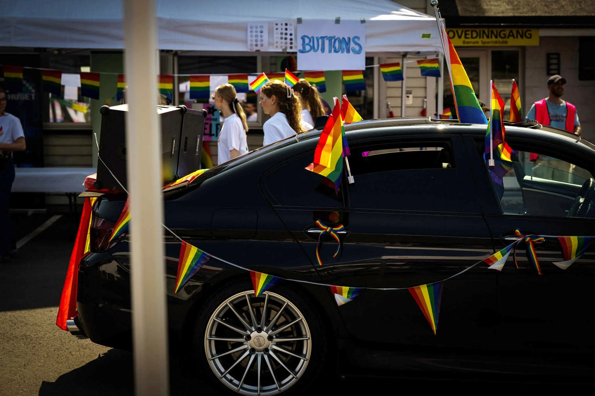 En svart bil dekorert med regnbuefargede flagg og bannere, parkert ved en Pride-parade med mennesker som går i bakgrunnen, og en skilt som sier 'BUTTONS'.