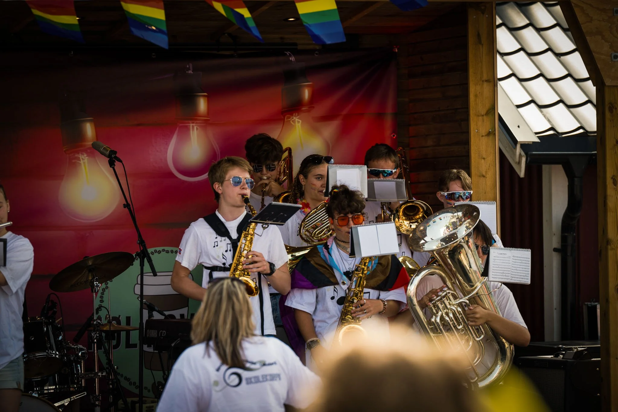 En gruppe unge musikere spiller på instrumenter som saksofon, tuba og trommer under en utendørs konsert, de bruker solbriller og står foran en bakgrunn med bilder av lyspærer.