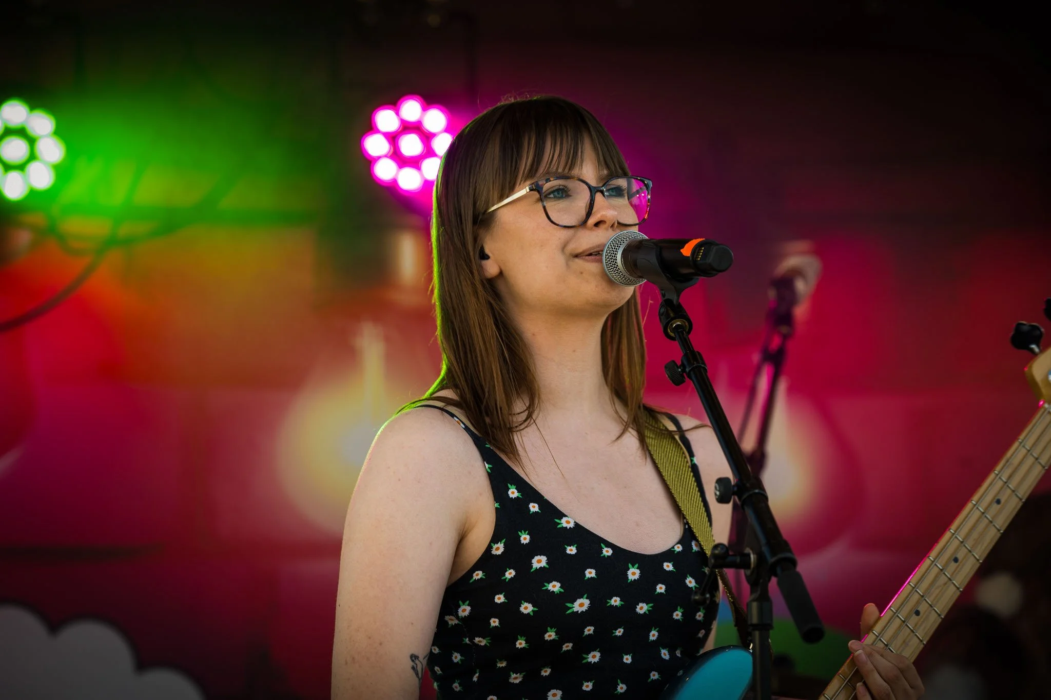 En kvinnelig musiker med briller og tatovering spiller gitar og synger inn i mikrofon på scenen med fargerikt belysning bak henne.