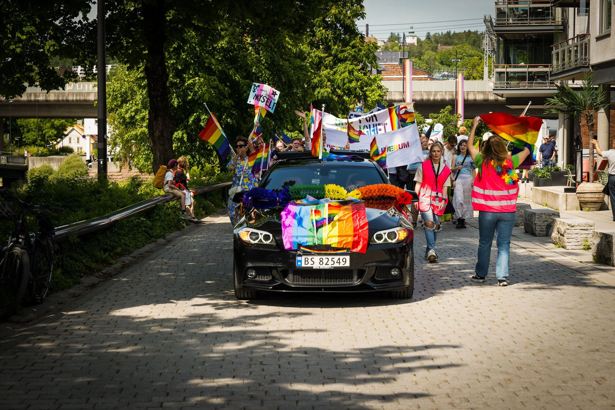 En gruppe mennesker deltar i en pride-parade, bærer regnbuefargede flag og banner, og går langs en gate med pyntede biler og trær.