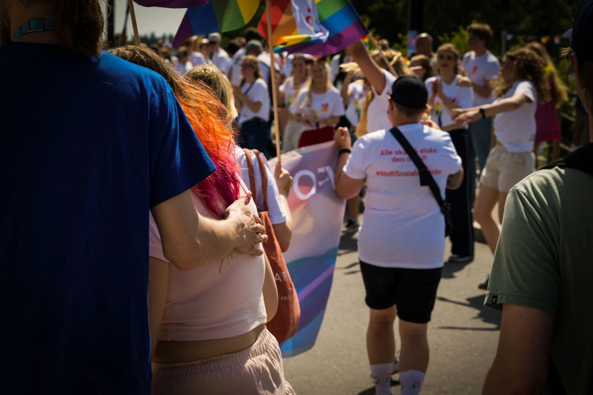 Folk som deltar i en parade, mange med regnbuefargede paraplyer og oppvisning, utendørs på en solskinnsdag.