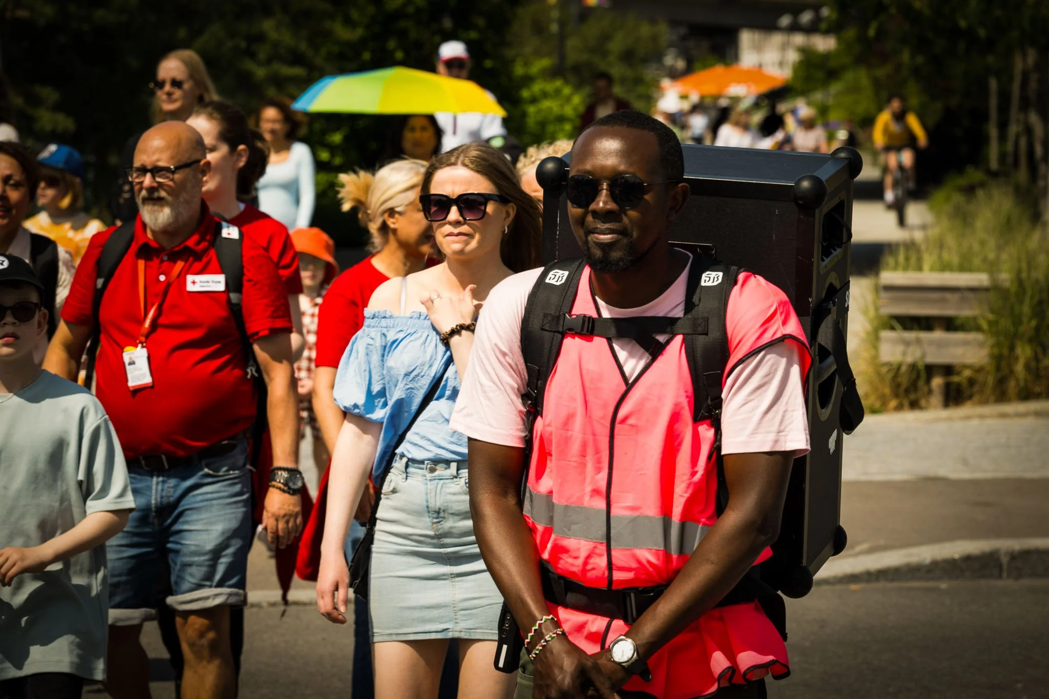 En gruppe mennesker går i solskinn, noen holder lyse paraplyer, en person bærer en stor bærbar høyttaler på ryggen, og flere går på en vei omgitt av grønt landskap