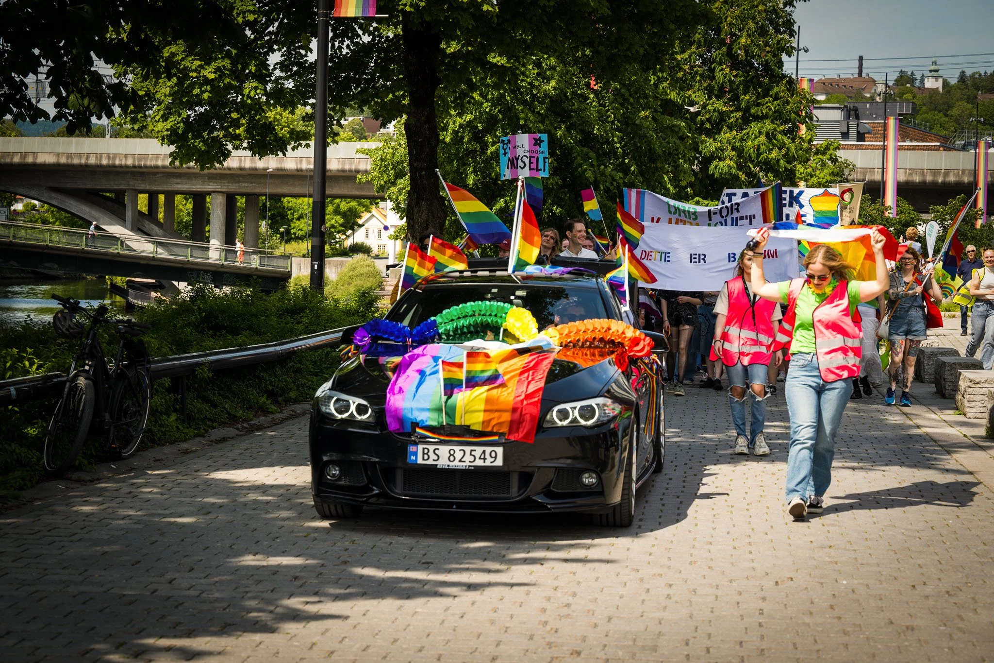 En svart bil pyntet med regnbuefargede pynt, kjører i en parade eller feiring med mennesker som bærer regnbuefargede flag og banner. Mange av deltakerne har på seg rosa vester og bærer regnbuefargede symboler, antageligvis for å feire Pride.
