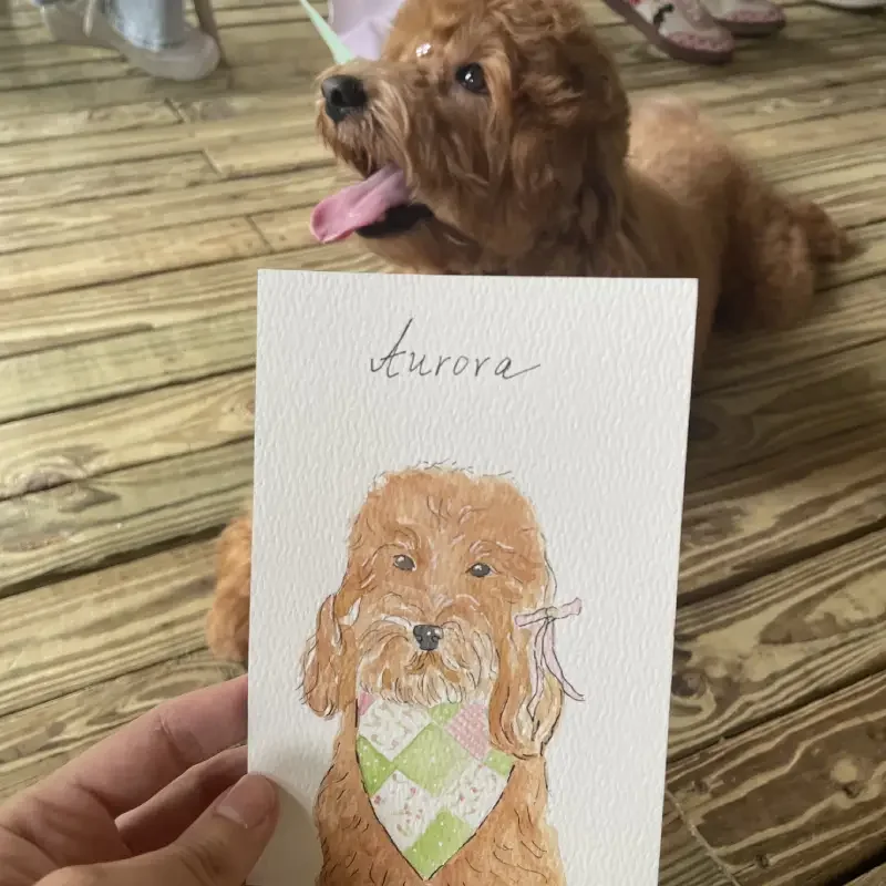 A sketchbook showing a drawing of a beach scene with a woman lying on a towel, trees, and the ocean, held against a real beach background with blue sky and some trees.