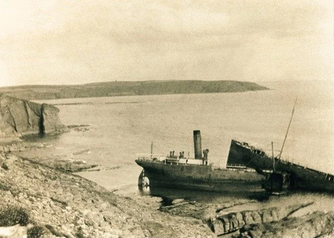 Historical black-and-white photo of a shipwreck on a rocky shore with cliffs in the background.