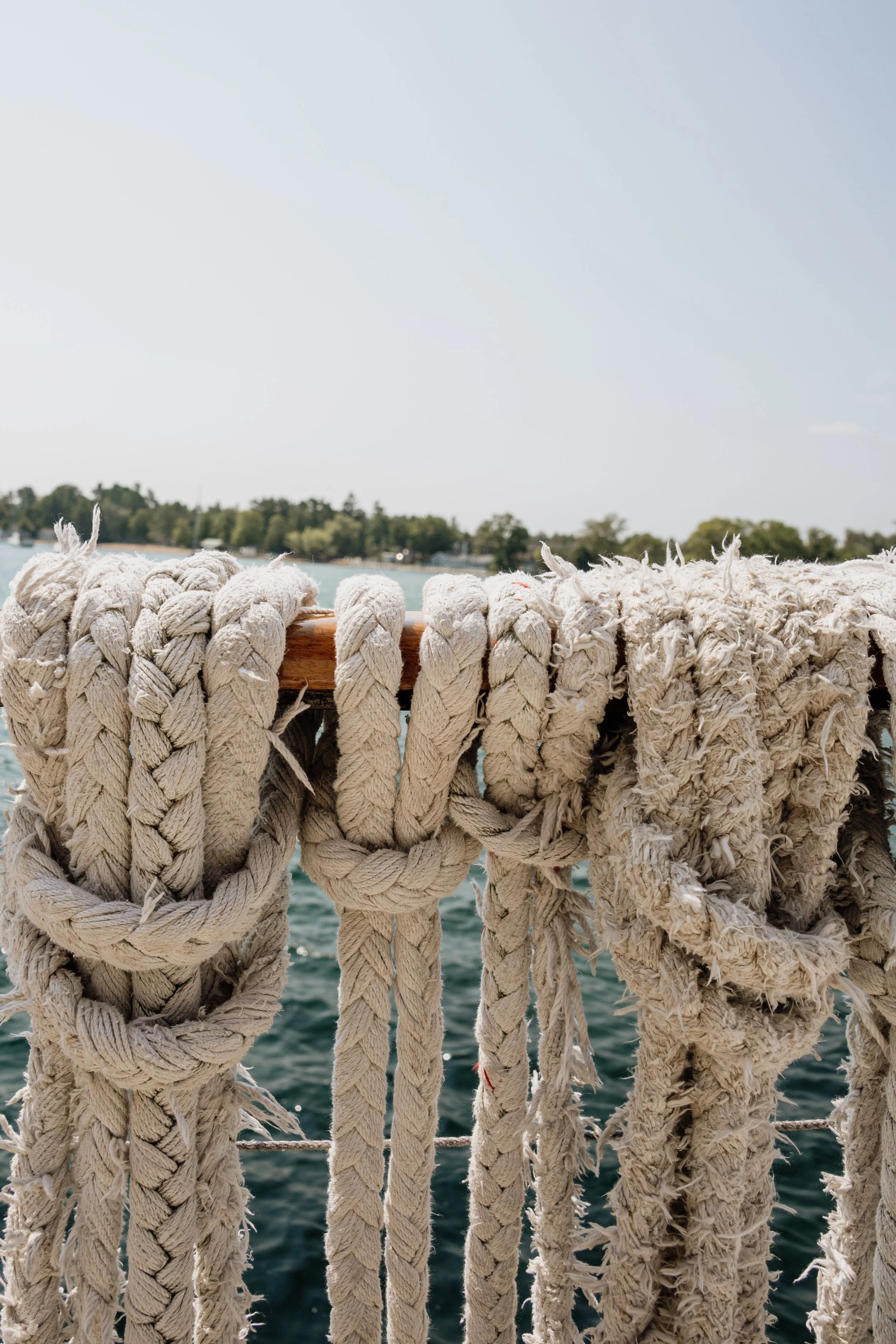 Close-up of weathered white ropes tied around a wooden railing on a boat, with a blurred body of water and a distant shoreline in the background under a clear sky.