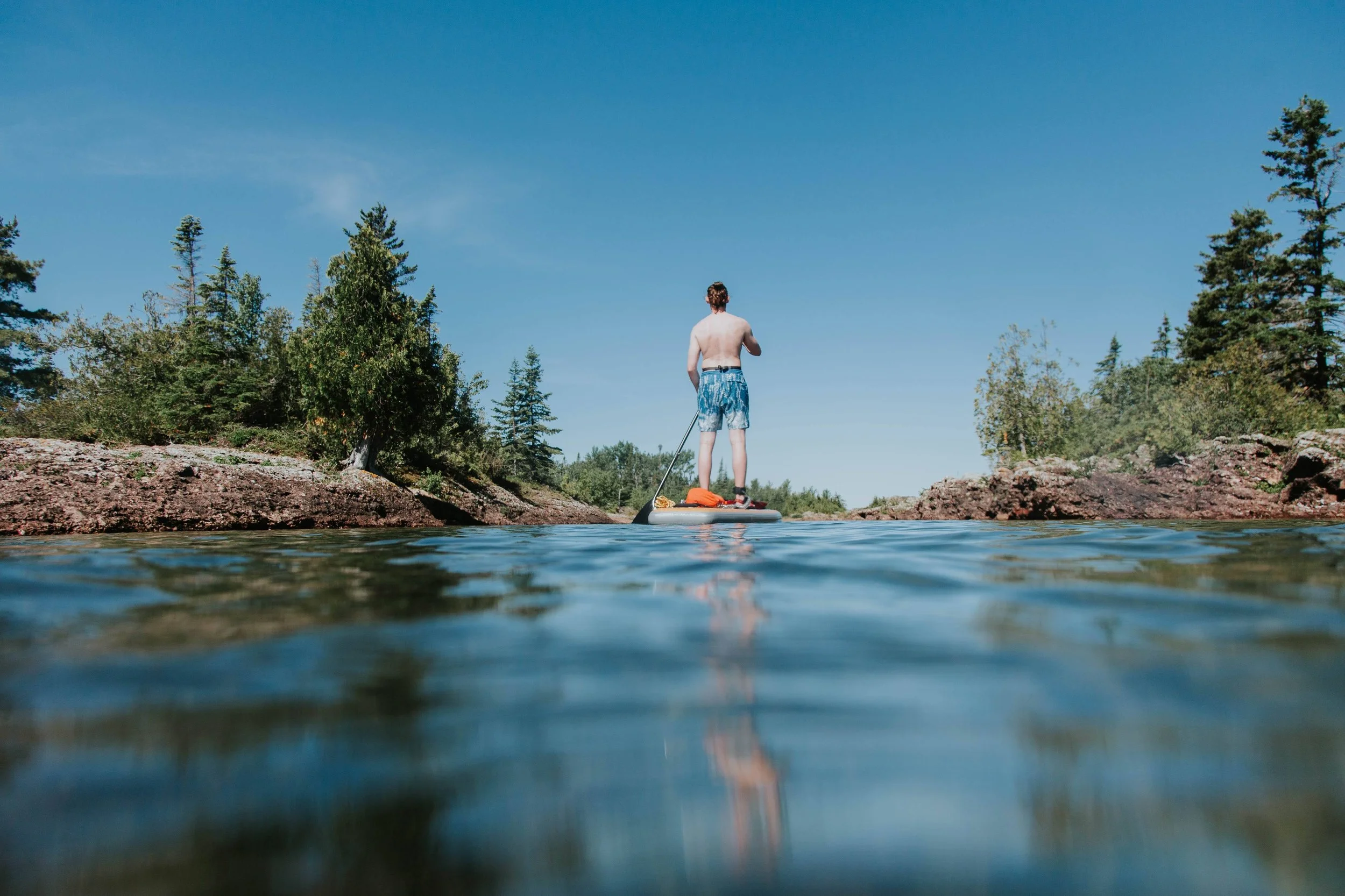 A man standing on an inflatable paddleboard in a river, surrounded by trees and rocks under a clear blue sky.